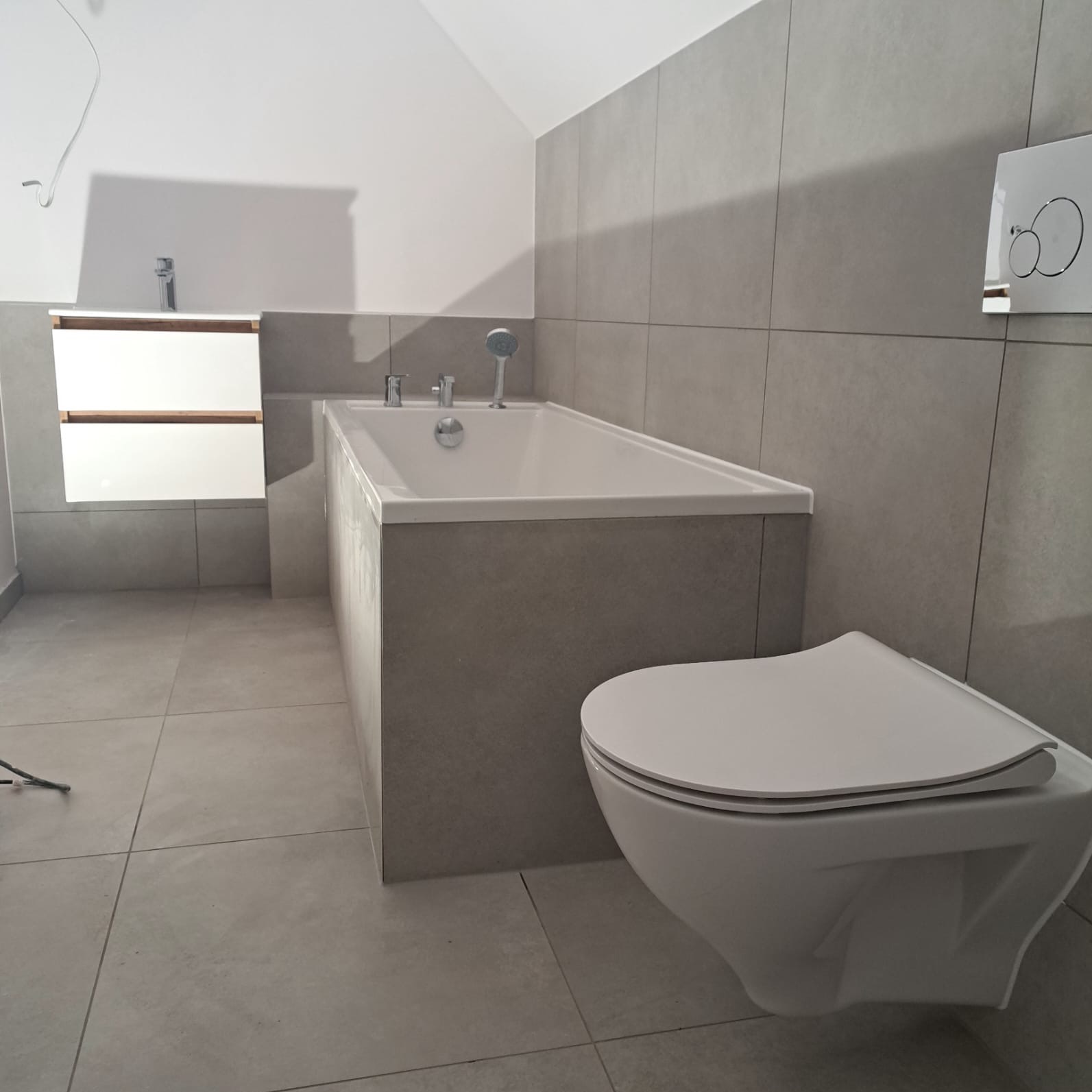 Nowoczesna łazienka w szarościach z podwieszaną miską WC, zabudowaną wanną z baterią i szafką pod umywalką, wykończona płytkami ceramicznymi.