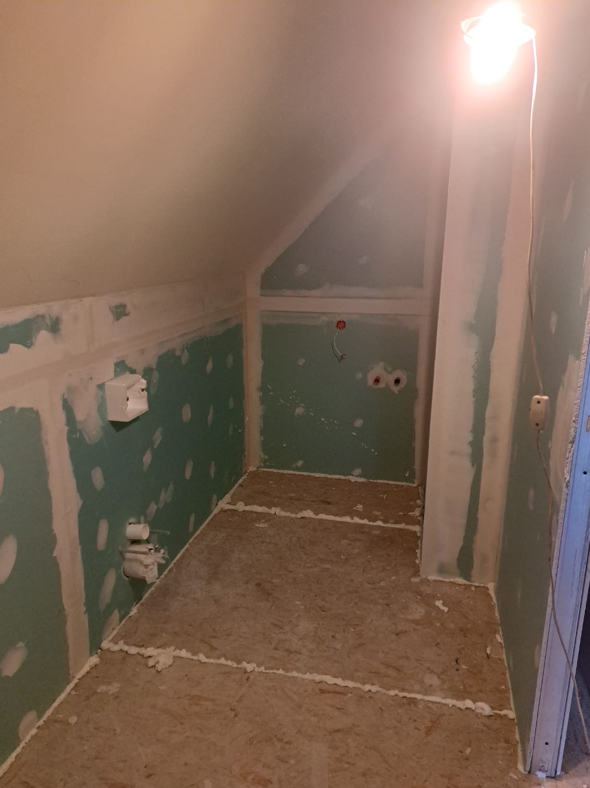 Poddasze w trakcie adaptacji: płyty gipsowo-kartonowe na ścianach, niedokończone szpachlowanie, podłoga z płyt OSB z widoczną pianką montażową, tymczasowe oświetlenie halogenowe.