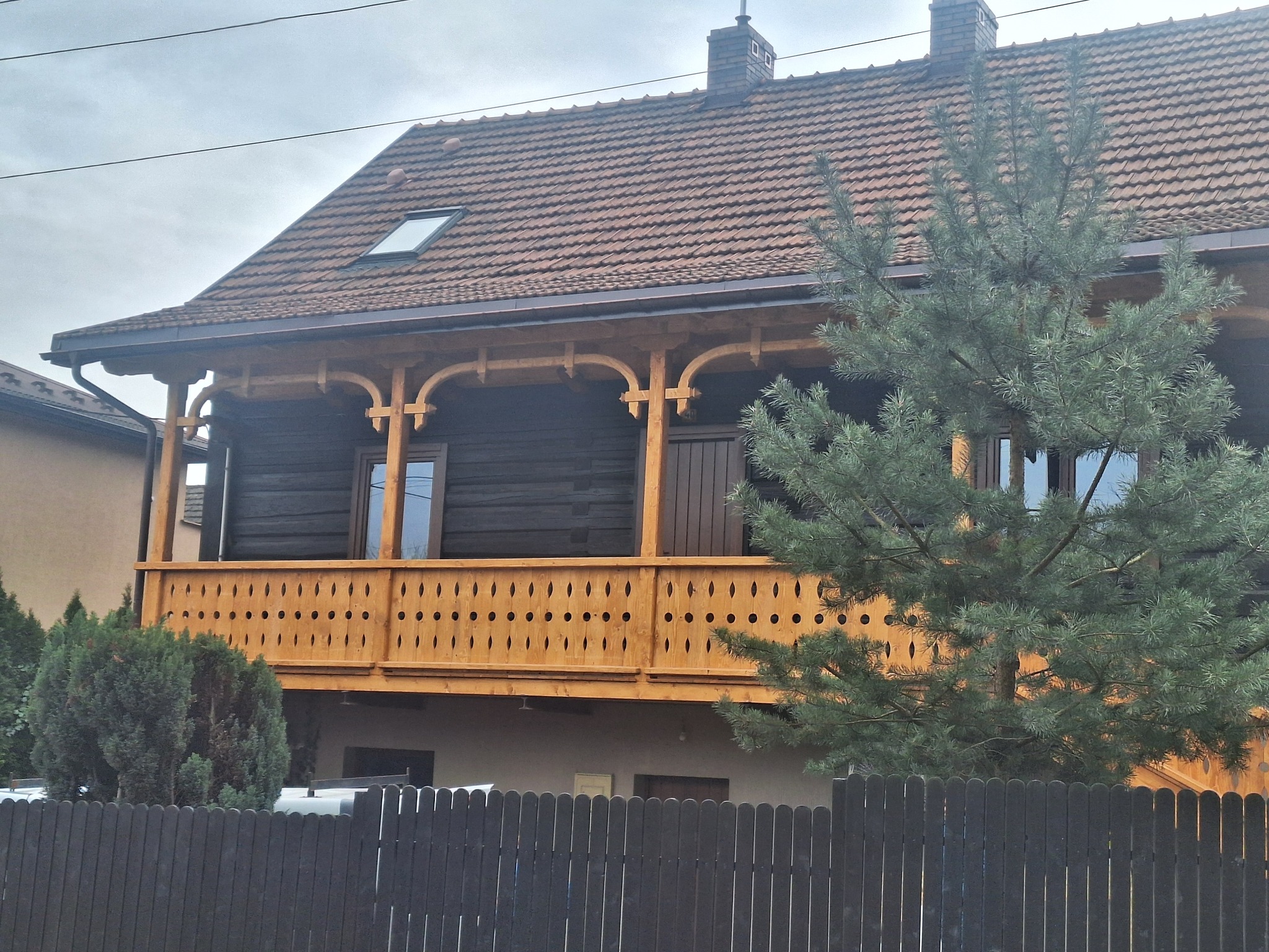 Dwukondygnacyjny dom z bali z drewnianym balkonem i brązowym dachem z lukarną, częściowo zasłonięty przez iglaste drzewo i ciemne ogrodzenie.