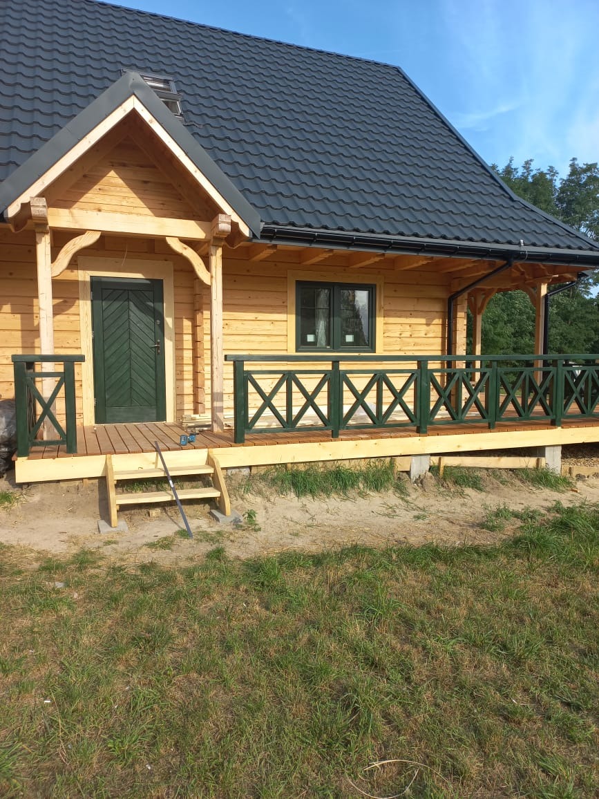 Drewniany dom z bali z ciemnym dachem i zielonymi akcentami. Widoczny taras z balustradą i schody wejściowe. Ujęcie w słoneczny dzień.