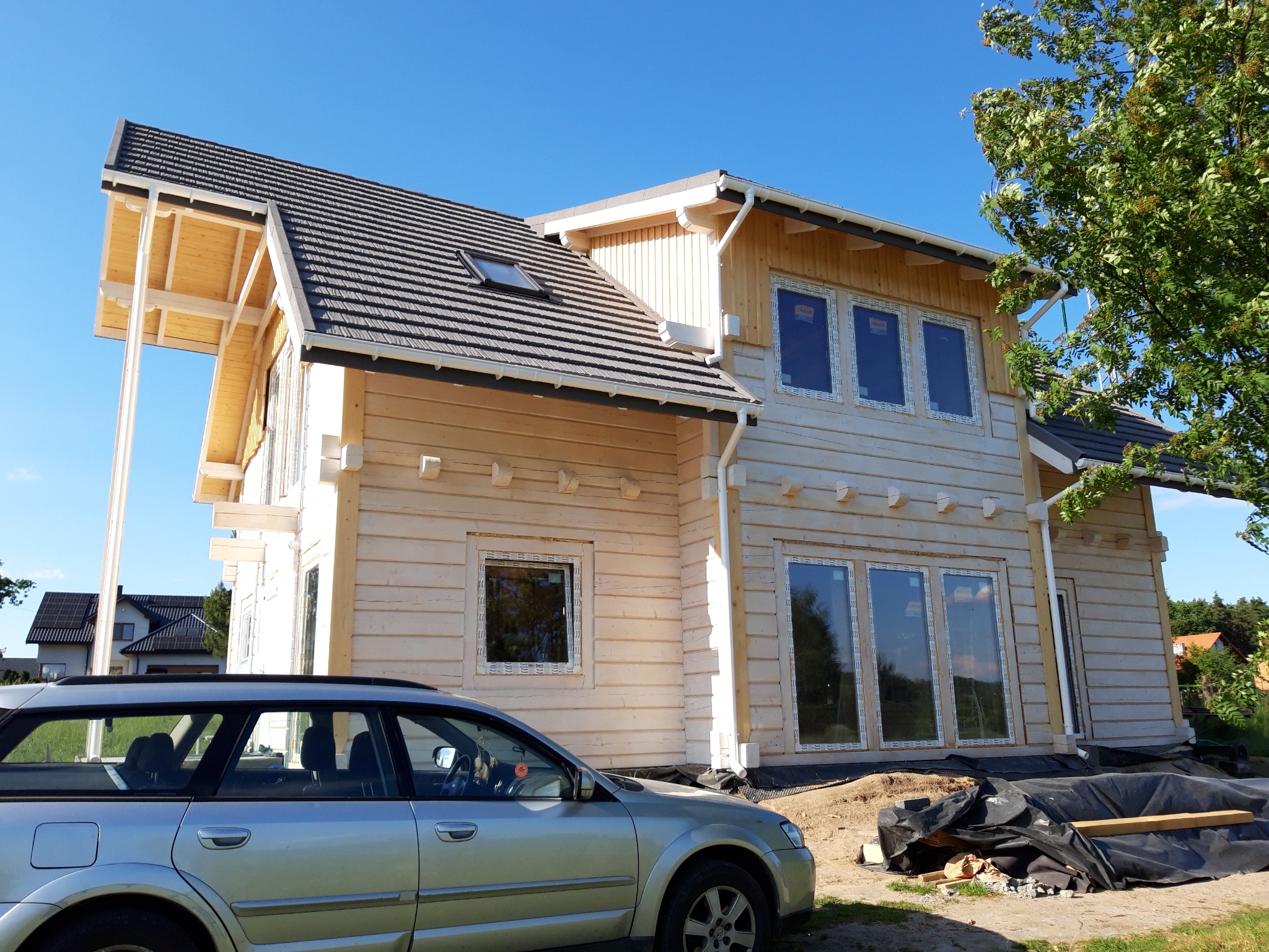 Surowy dom z bali w trakcie budowy, z widoczną konstrukcją dachu i oknami, na tle błękitnego nieba i zieleni. Obok zaparkowany srebrny samochód.