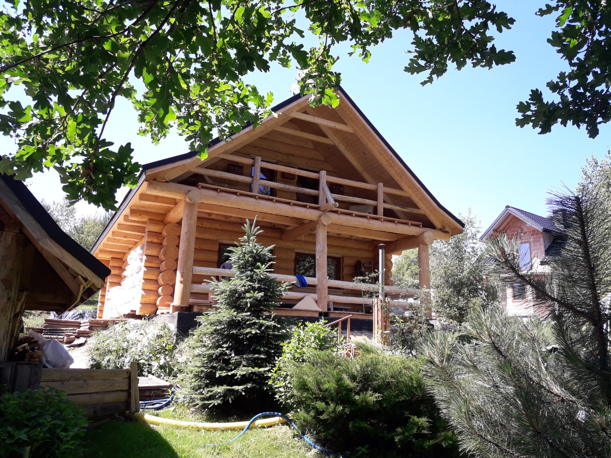 Dwukondygnacyjny dom z bali, z balkonem i widoczną konstrukcją dachu, otoczony zielenią i drzewami, w słoneczny dzień. Widok z poziomu gruntu.