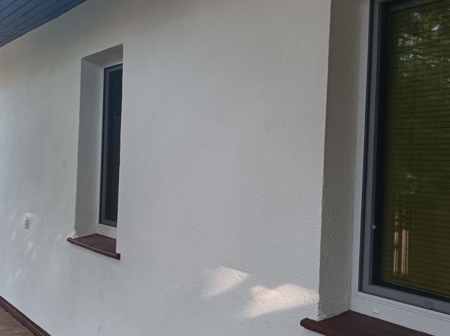 Biała, teksturowana ściana zewnętrzna budynku z dwoma oknami o szarych ramach i brązowymi parapetami, oświetlona naturalnym światłem słonecznym.