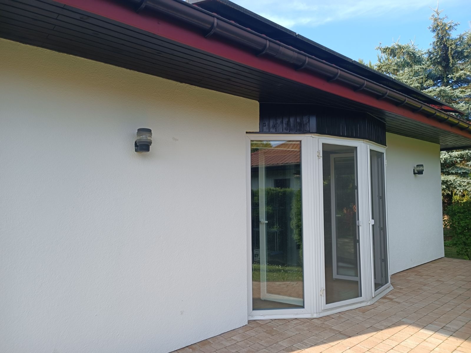 Elewacja domu z białym tynkiem, ciemnym dachem z rynnami i narożnym, przeszklonym wejściem z białymi ramami okiennymi.