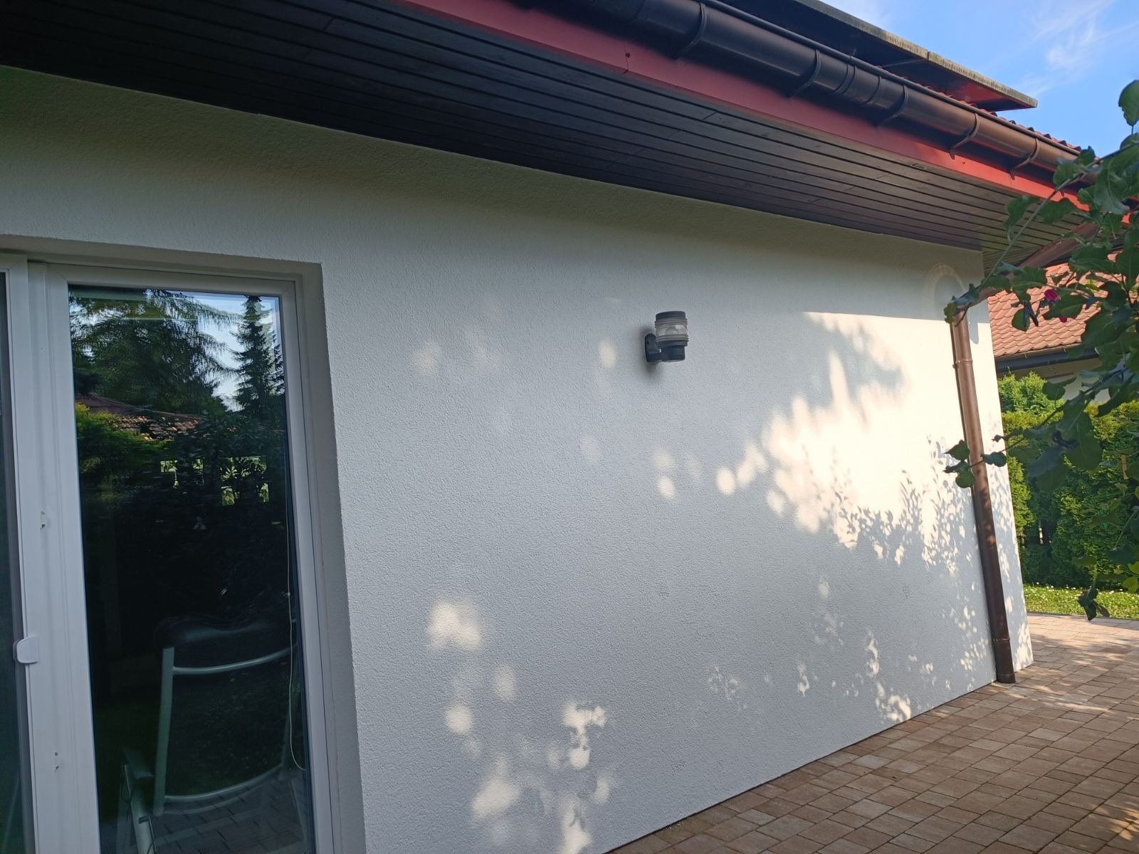 Biała, świeżo otynkowana ściana zewnętrzna domu z zamontowaną lampą, widoczny fragment okna i brązowa rynna, na kostce brukowej padają cienie drzew.