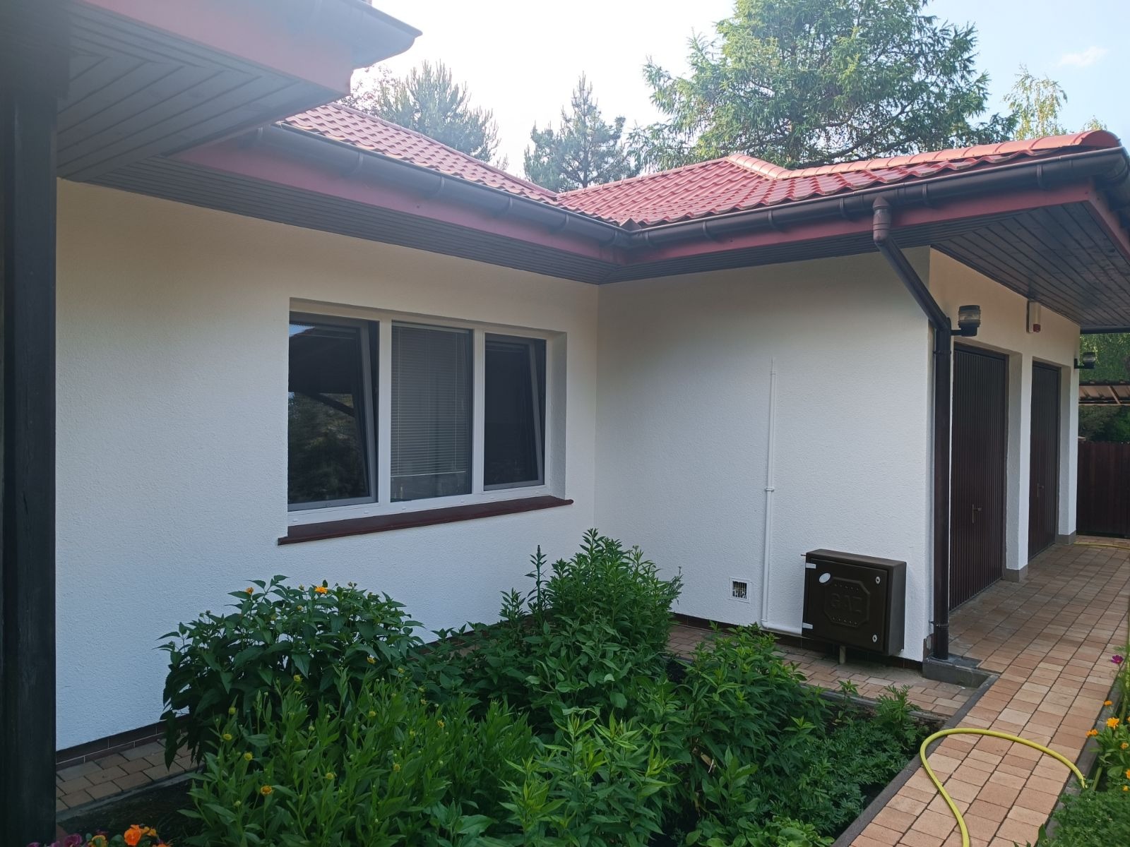 Świeżo pomalowana elewacja domu z czerwonym dachem, widoczne okno z żaluzjami, zadbany ogródek i brązowe drzwi garażowe.