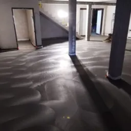 Świeżo wylana, szara posadzka z widocznymi śladami po zacieraczce, w surowym wnętrzu z betonowymi filarami i pracującym mężczyzną w czerwonej czapce w tle.