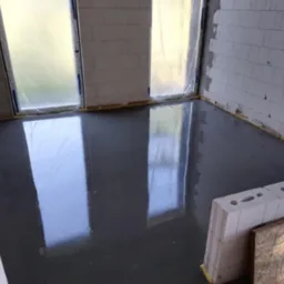 Świeżo wylana, gładka posadzka przemysłowa w surowym wnętrzu z białych bloczków, zabezpieczone okna folią. Widoczne odbicia światła na mokrej powierzchni.