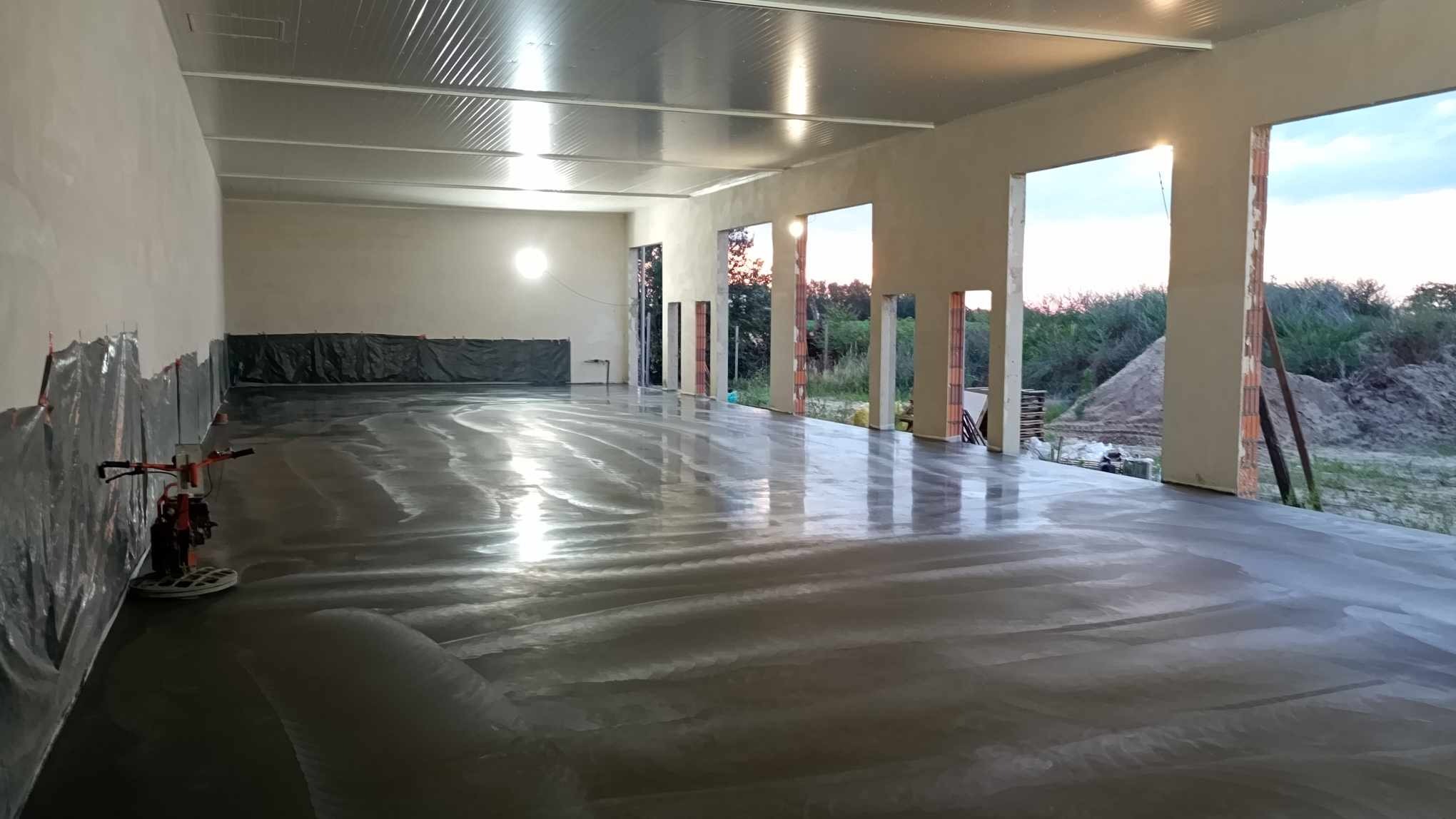 Świeżo wylana, mokra posadzka w hali przemysłowej, zabezpieczona folią przy ścianach, zacierana mechanicznie; widoczne otwarte okna z widokiem na zewnątrz.