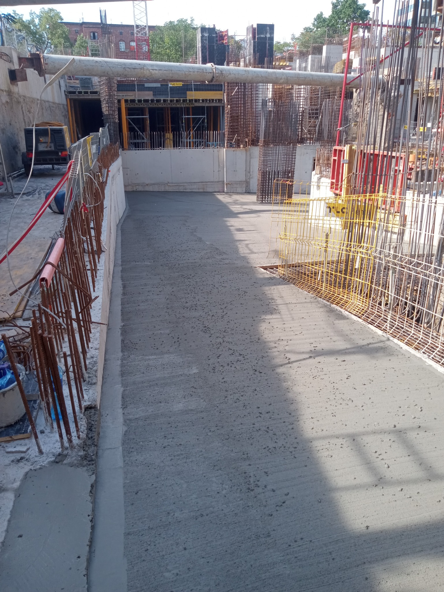 Świeżo wylana, równa powierzchnia betonowa na placu budowy, otoczona zbrojeniami i szalunkami, z widocznym sprzętem budowlanym w tle.