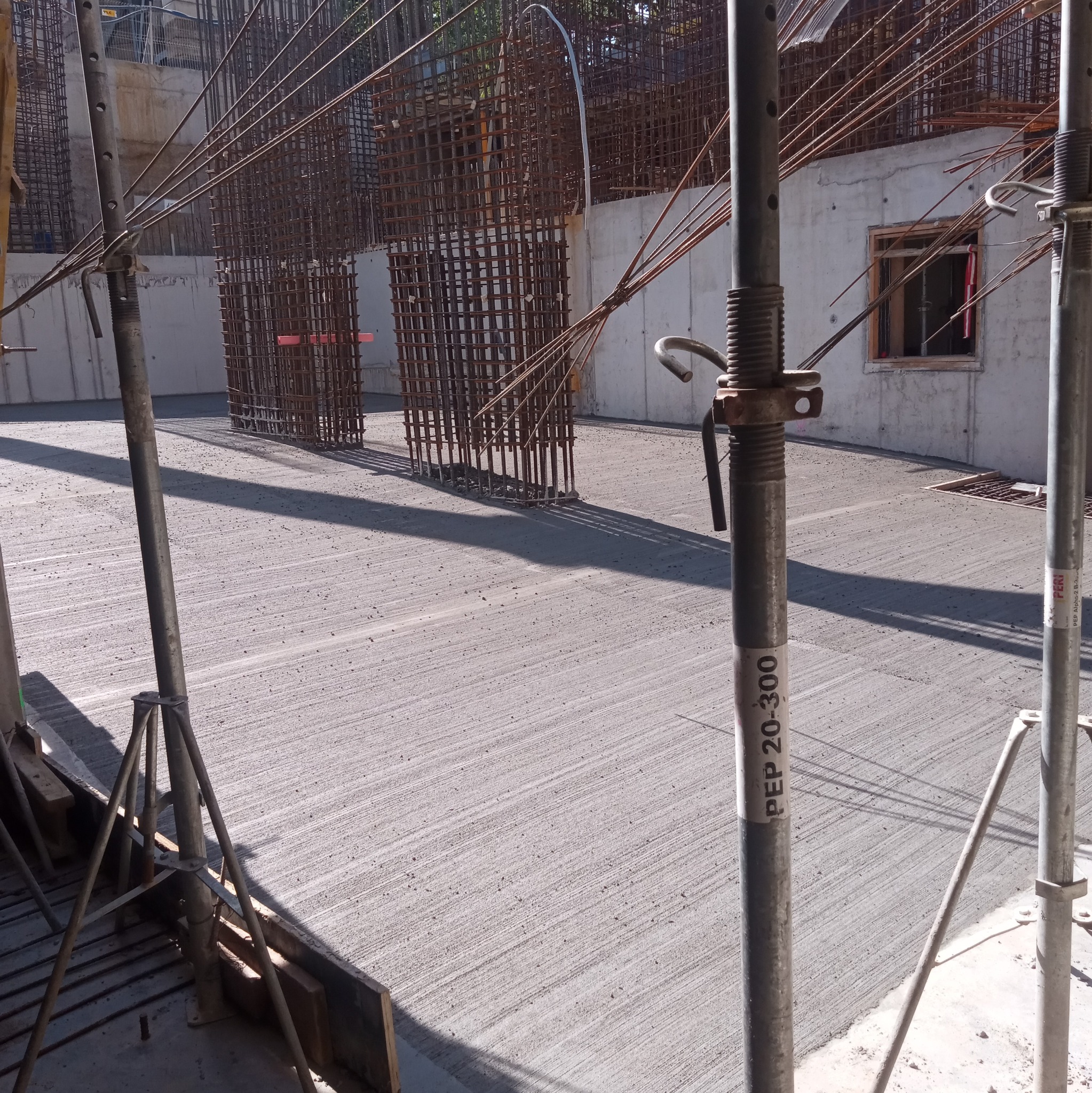 Świeżo wylana, szara betonowa posadzka przemysłowa z widocznymi śladami po zacieraniu, otoczona metalowymi szalunkami i podporami, zbrojeniami w tle i fragmentem ściany z oknem.