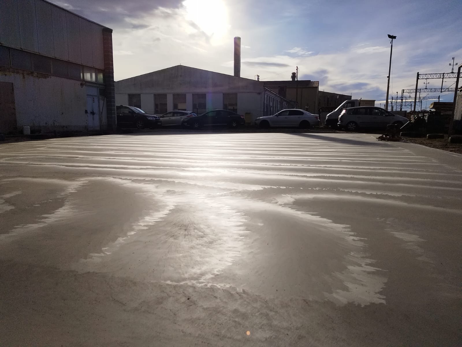 Świeżo wylana, mokra posadzka przemysłowa na zewnątrz budynku, z widocznymi śladami po wyrównywaniu i odbiciami nieba; w tle zaparkowane samochody i budynki przemysłowe.