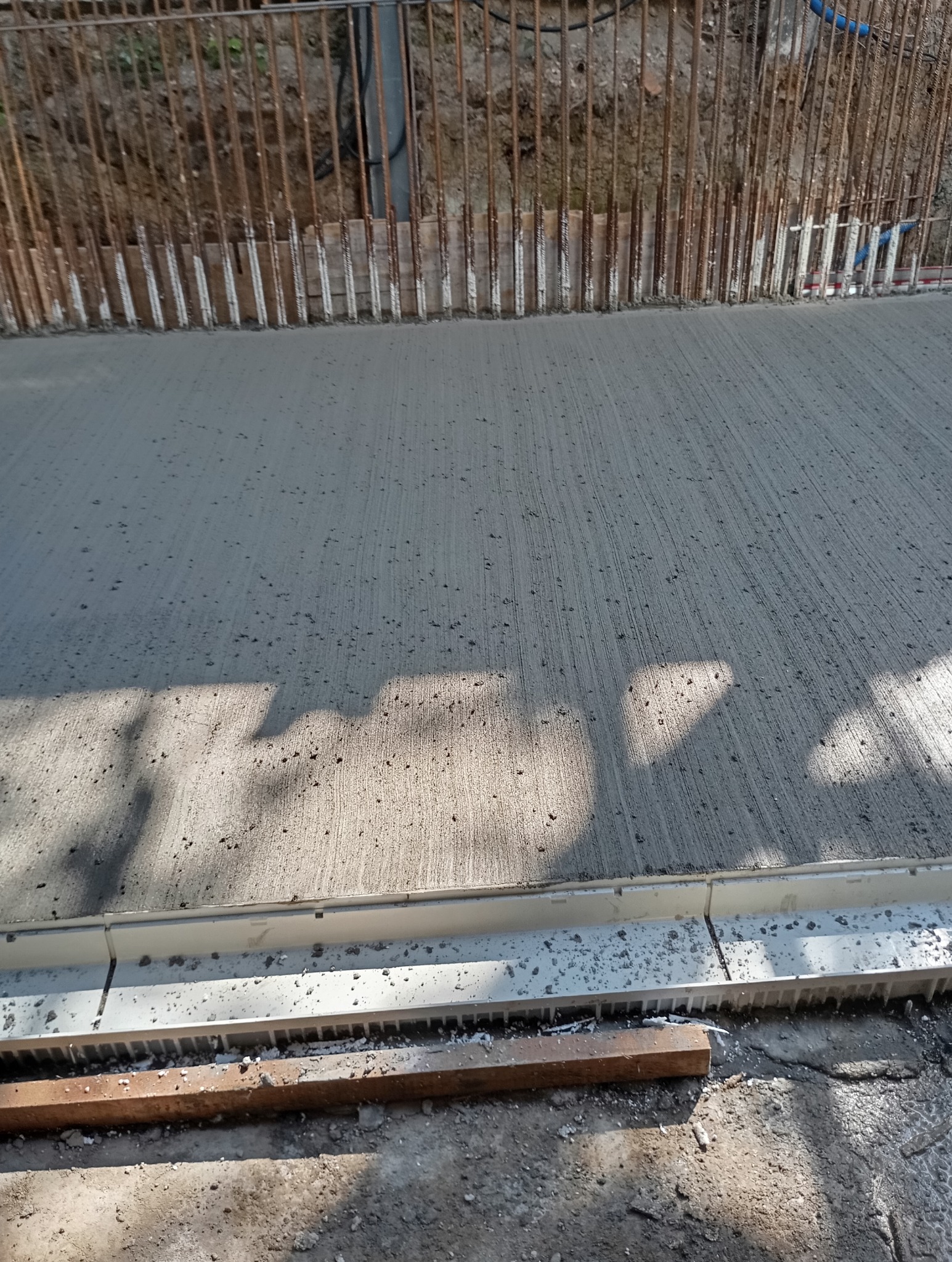 Świeżo wylana, szara powierzchnia z widocznymi śladami po zacieraniu, ograniczona szalunkiem i zbrojeniem pionowym w tle. Fragmenty cienia na powierzchni.