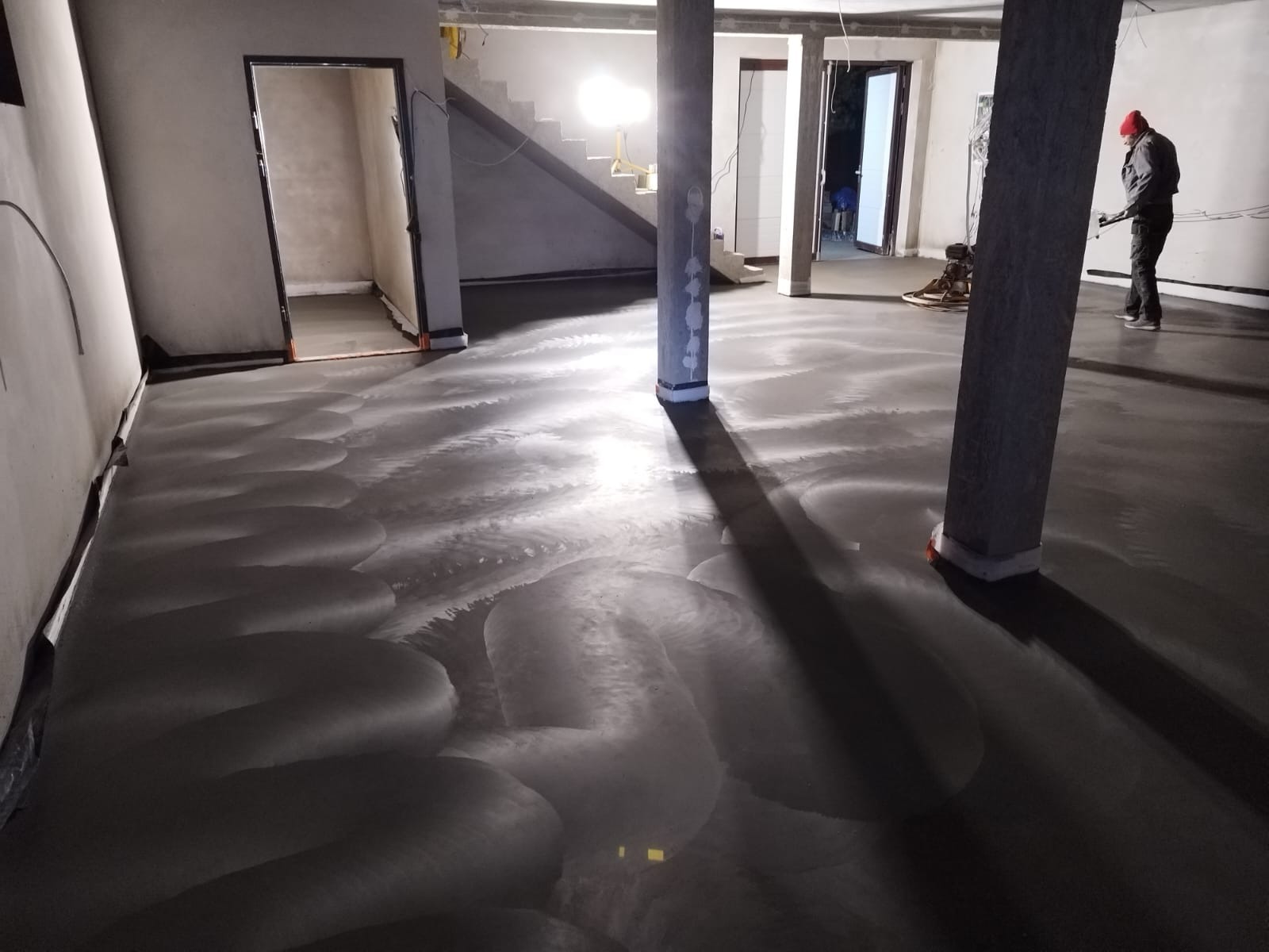 Świeżo wylana, szara posadzka z widocznymi śladami po zacieraczce, w surowym wnętrzu z betonowymi filarami i pracującym mężczyzną w czerwonej czapce w tle.