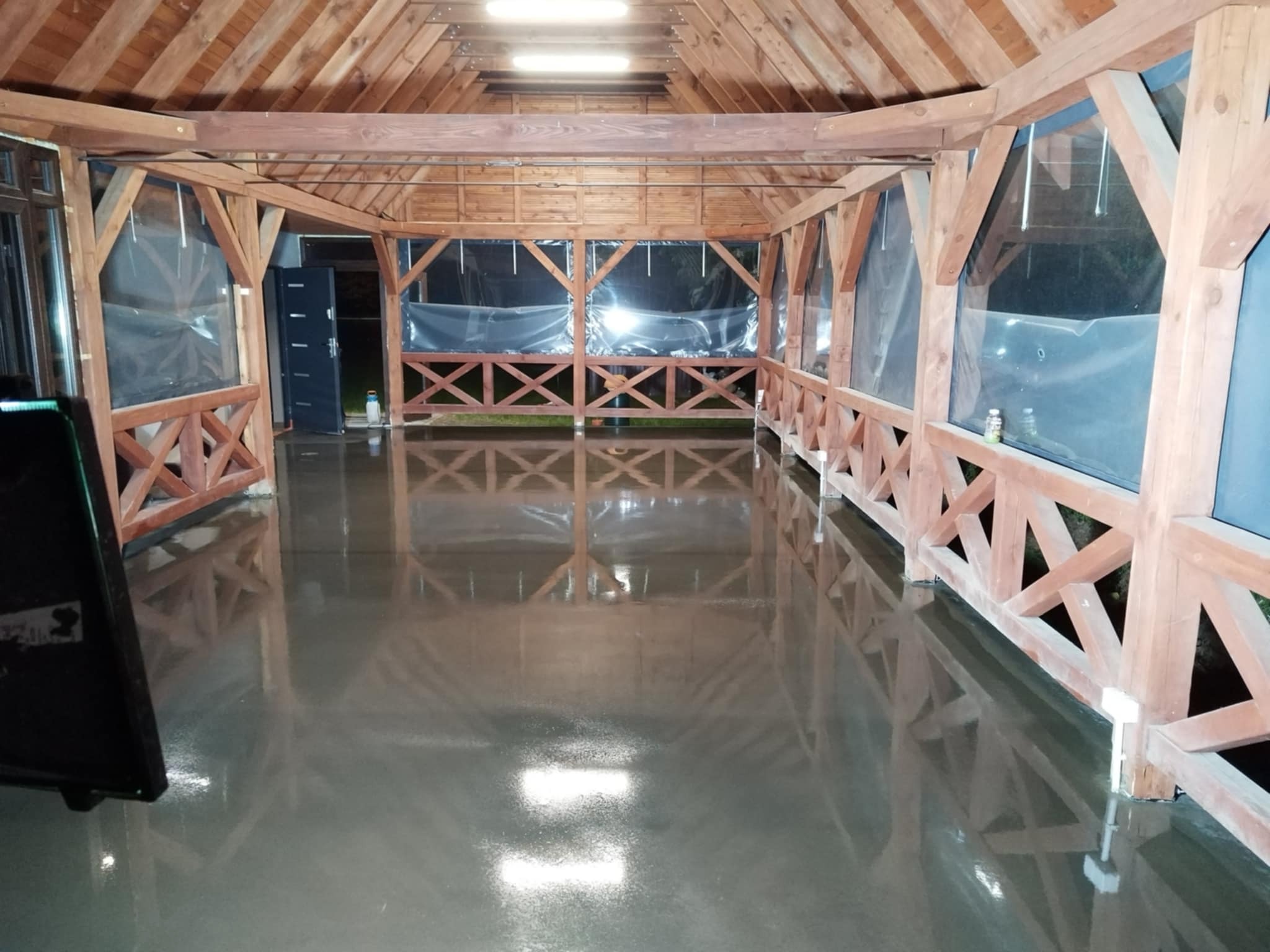 Świeżo wylana, błyszcząca posadzka przemysłowa w drewnianej altanie z przezroczystymi osłonami. Widoczne odbicia światła i drewniana konstrukcja.