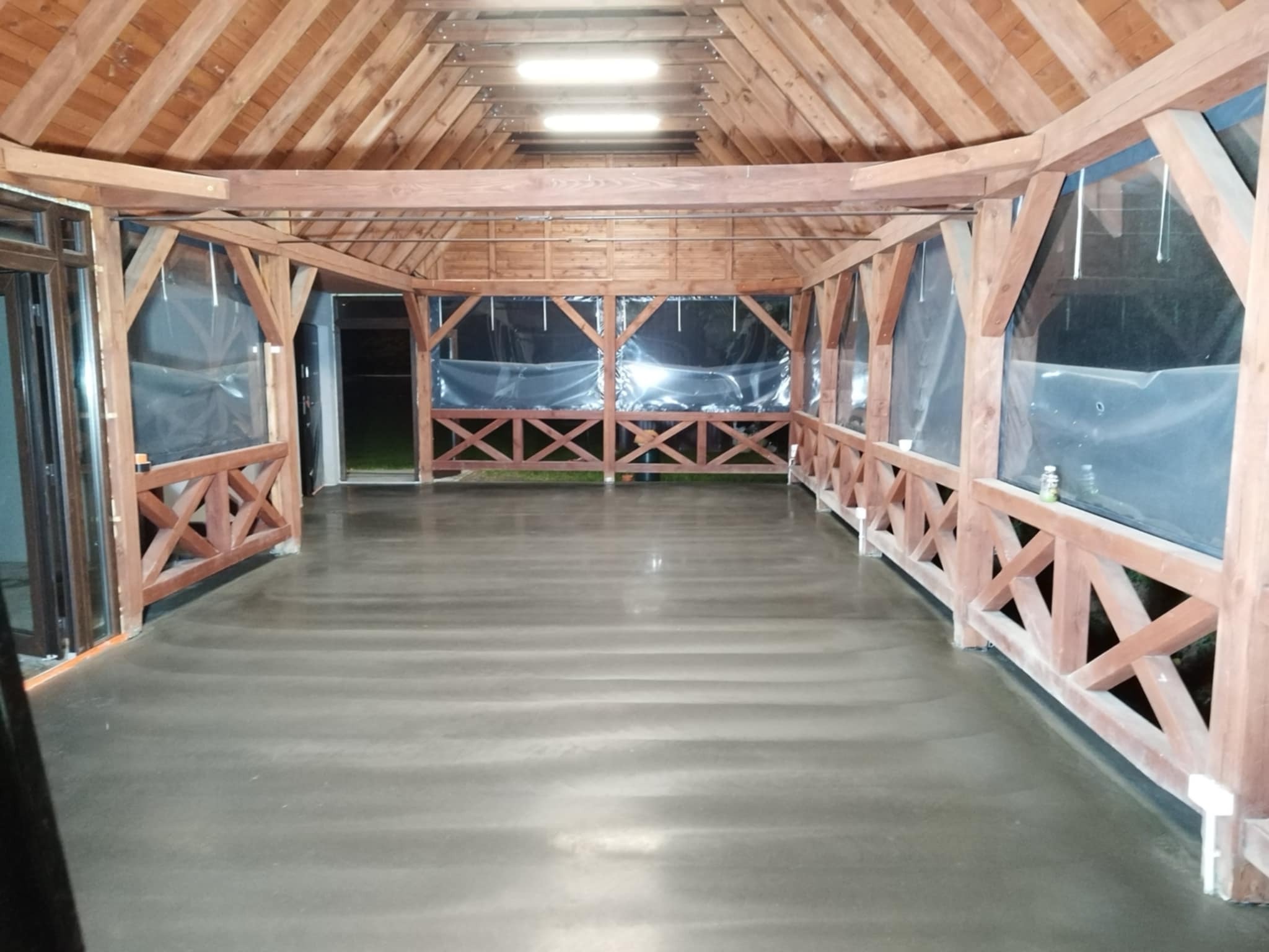 Wypolerowana, szara posadzka przemysłowa w drewnianej altanie z przezroczystymi zasłonami. Widoczne detale konstrukcyjne i oświetlenie.