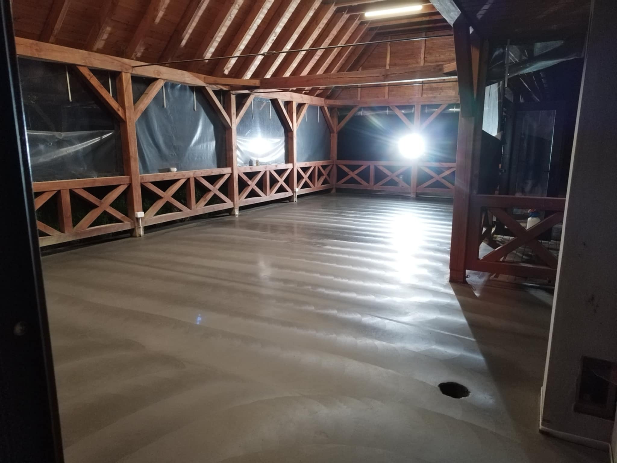 Świeżo wylana, gładka posadzka w drewnianej konstrukcji z zadaszeniem. Widoczne ślady po wyrównywaniu, otwór w podłodze. Oświetlenie sufitowe.