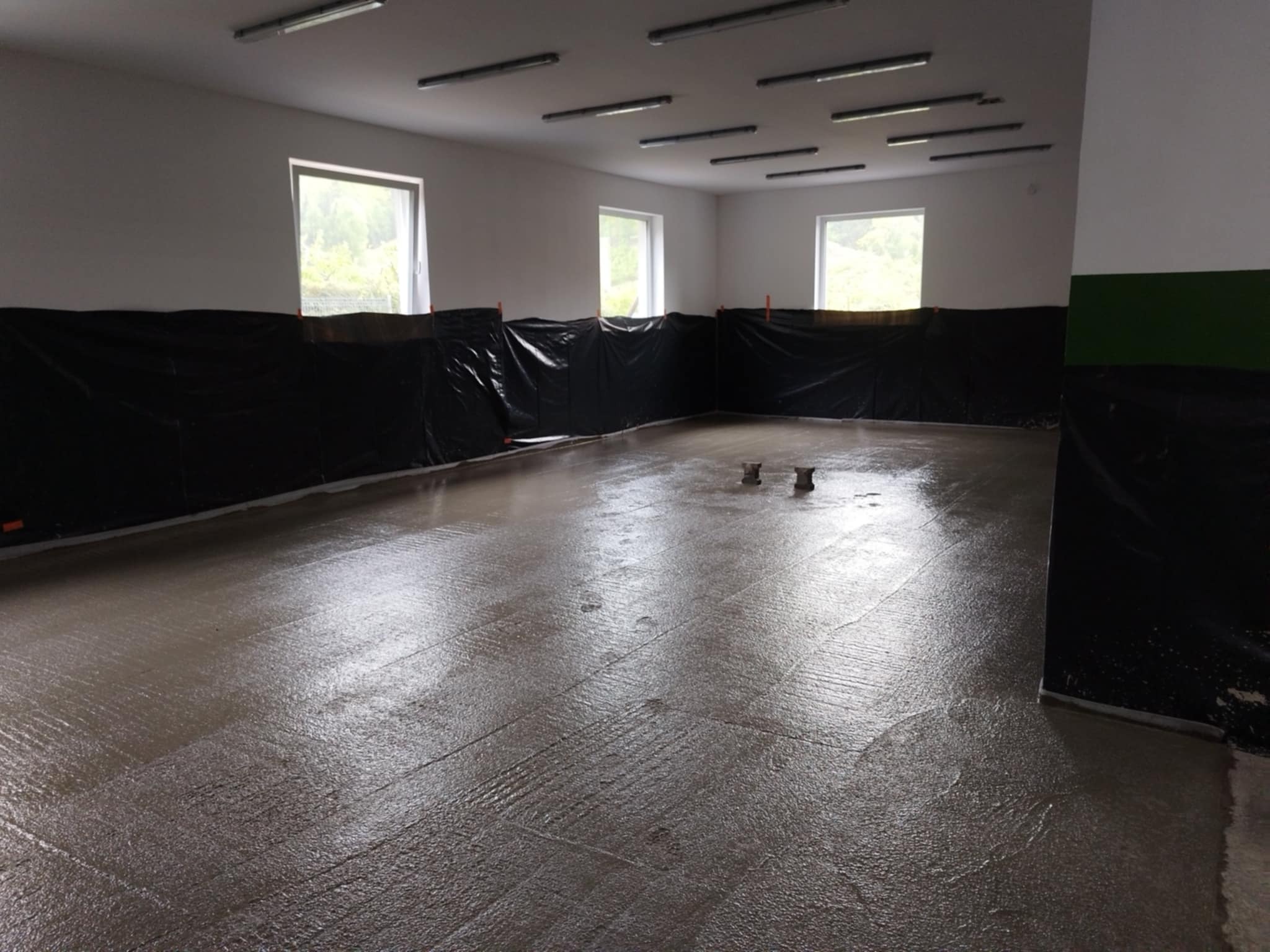 Wylana, świeża posadzka w dużym pomieszczeniu z oknami, zabezpieczonymi ścianami folią. Widoczne ślady po narzędziach i dwa wystające punkty w podłodze.