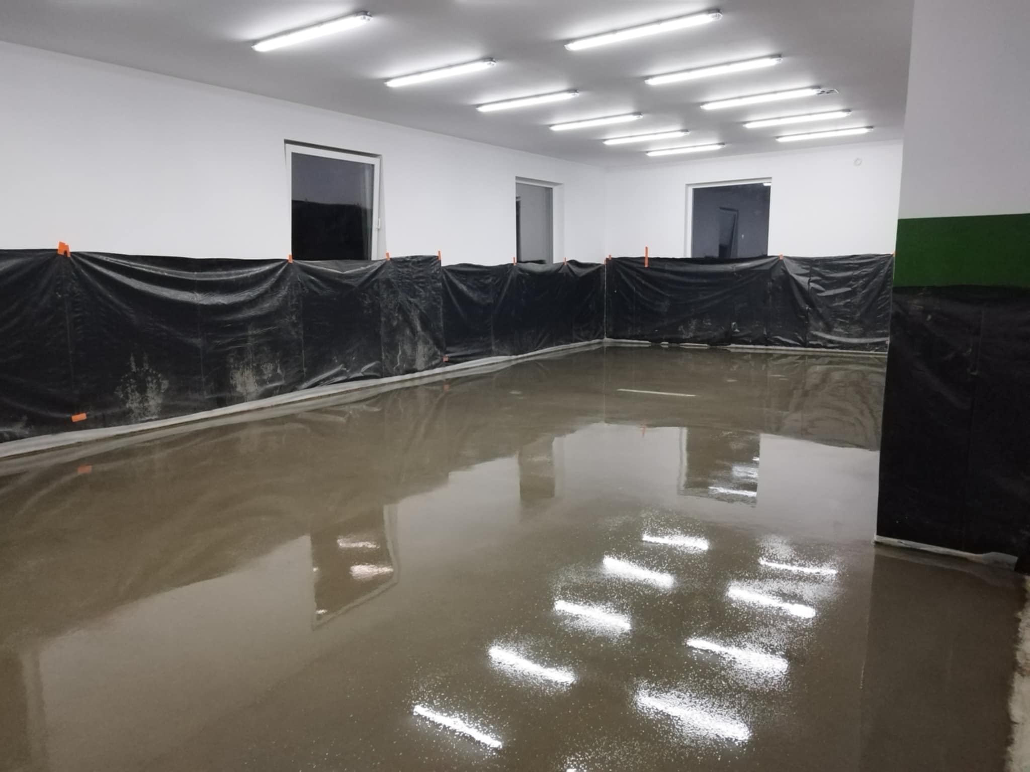 Świeżo wylana, błyszcząca posadzka przemysłowa w jasnym pomieszczeniu z oknami, zabezpieczonymi folią ochronną. Widoczne odbicia lamp sufitowych.