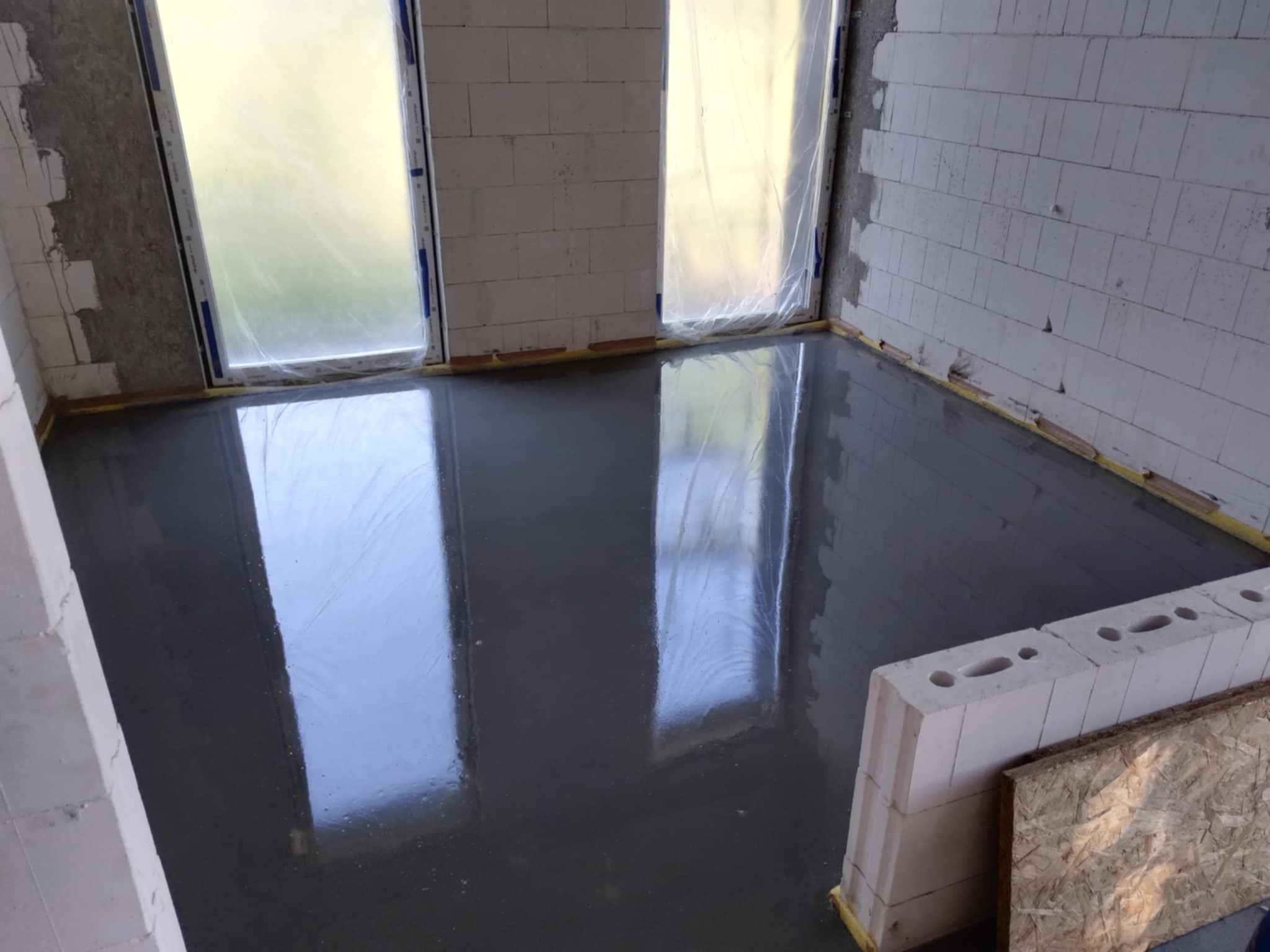 Świeżo wylana, gładka posadzka przemysłowa w surowym wnętrzu z białych bloczków, zabezpieczone okna folią. Widoczne odbicia światła na mokrej powierzchni.