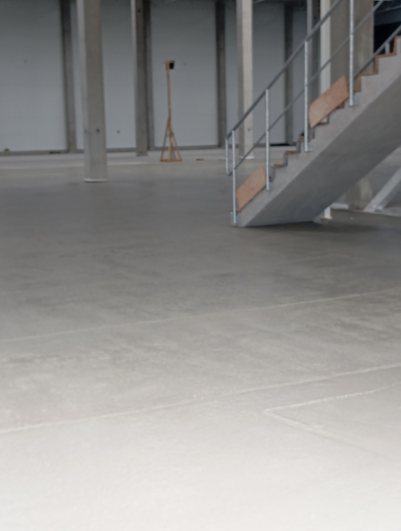 Jasnoszara, gładka posadzka przemysłowa w hali z betonowymi kolumnami i schodami. Widoczne ślady po szlifowaniu. Minimalistyczny, surowy wygląd.