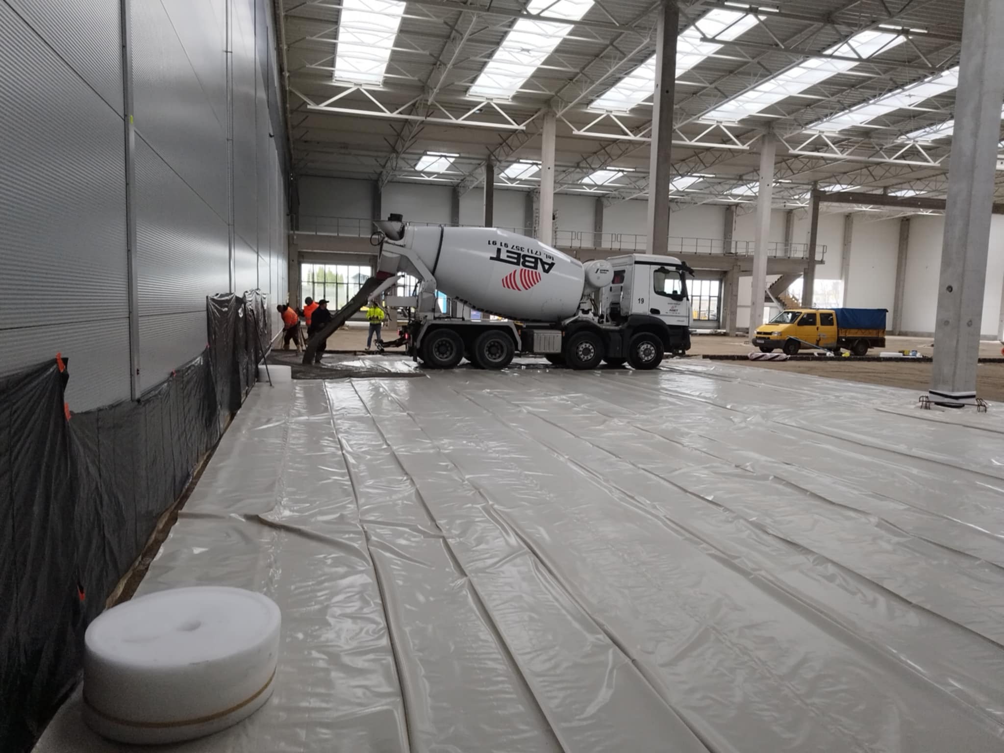 Wylewka betonu z betoniarki ABET na folii w hali przemysłowej. Widoczni pracownicy i żółty samochód dostawczy w tle. Wysoki sufit z oknami dachowymi.