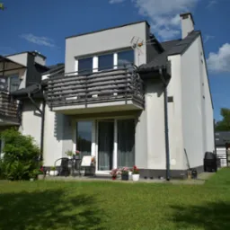 Dwupiętrowy dom szeregowy z balkonem z czarną balustradą i anteną na dachu, widok z ogrodu z trawnikiem, na balkonie stolik i krzesła oraz kwiaty w donicach.