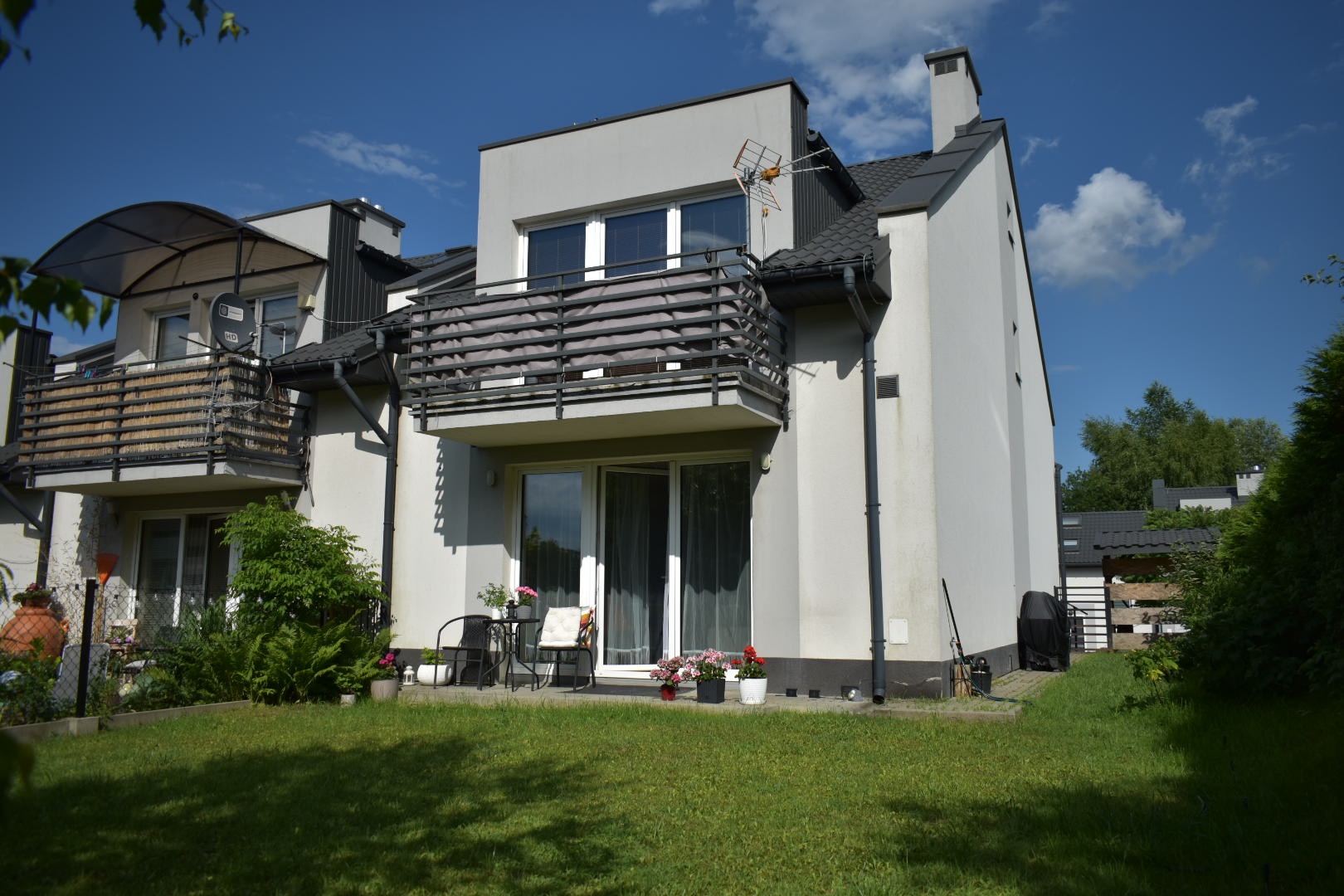 Dwupiętrowy dom szeregowy z balkonem z czarną balustradą i anteną na dachu, widok z ogrodu z trawnikiem, na balkonie stolik i krzesła oraz kwiaty w donicach.