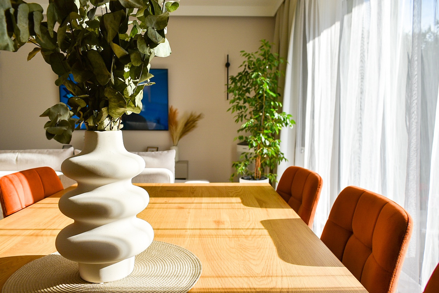Jasny salon z drewnianym stołem, pomarańczowymi krzesłami i dekoracyjnym wazonem z eukaliptusem w słonecznym świetle.