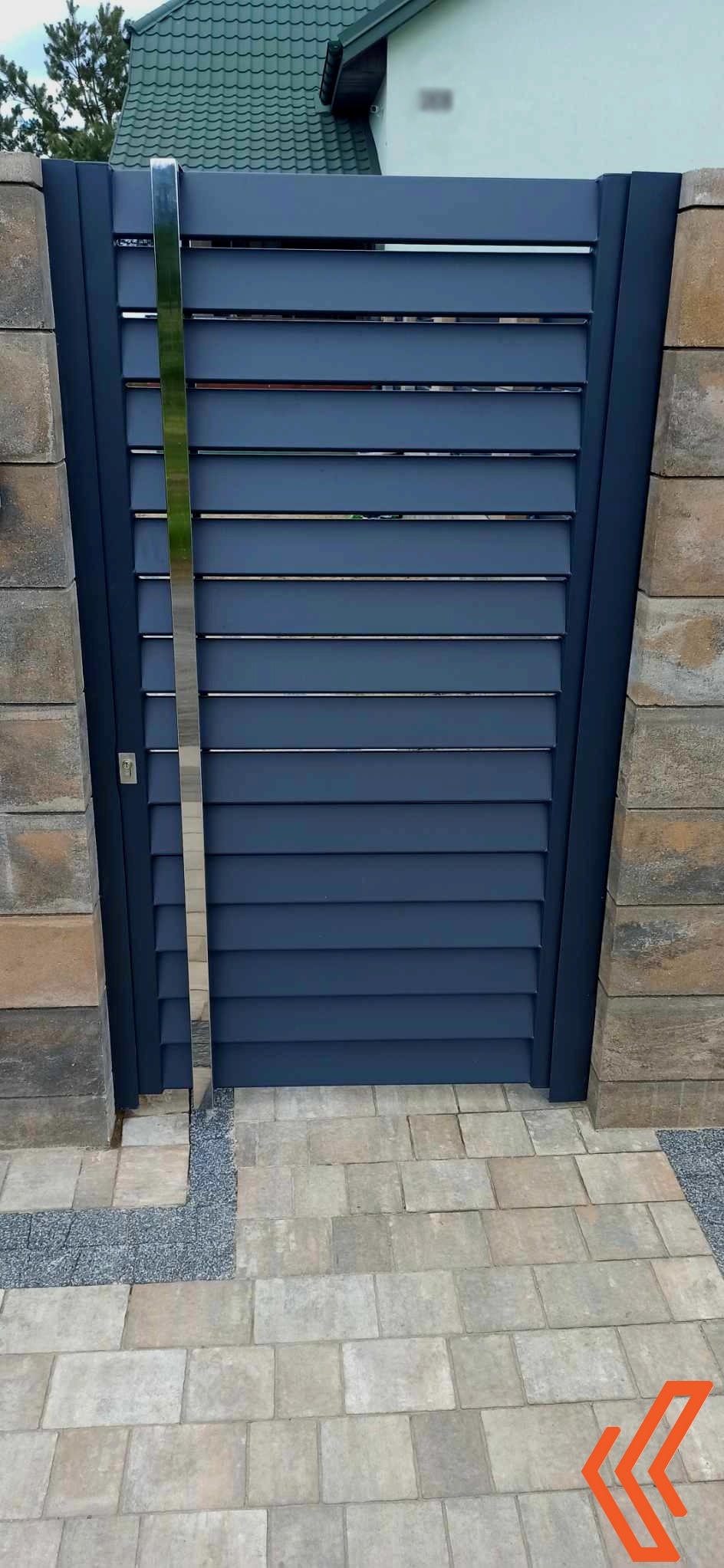 Nowoczesna brama wjazdowa z poziomych, szarych paneli, z błyszczącym, pionowym akcentem ze stali nierdzewnej, osadzona między kamiennymi słupkami na kostce brukowej.