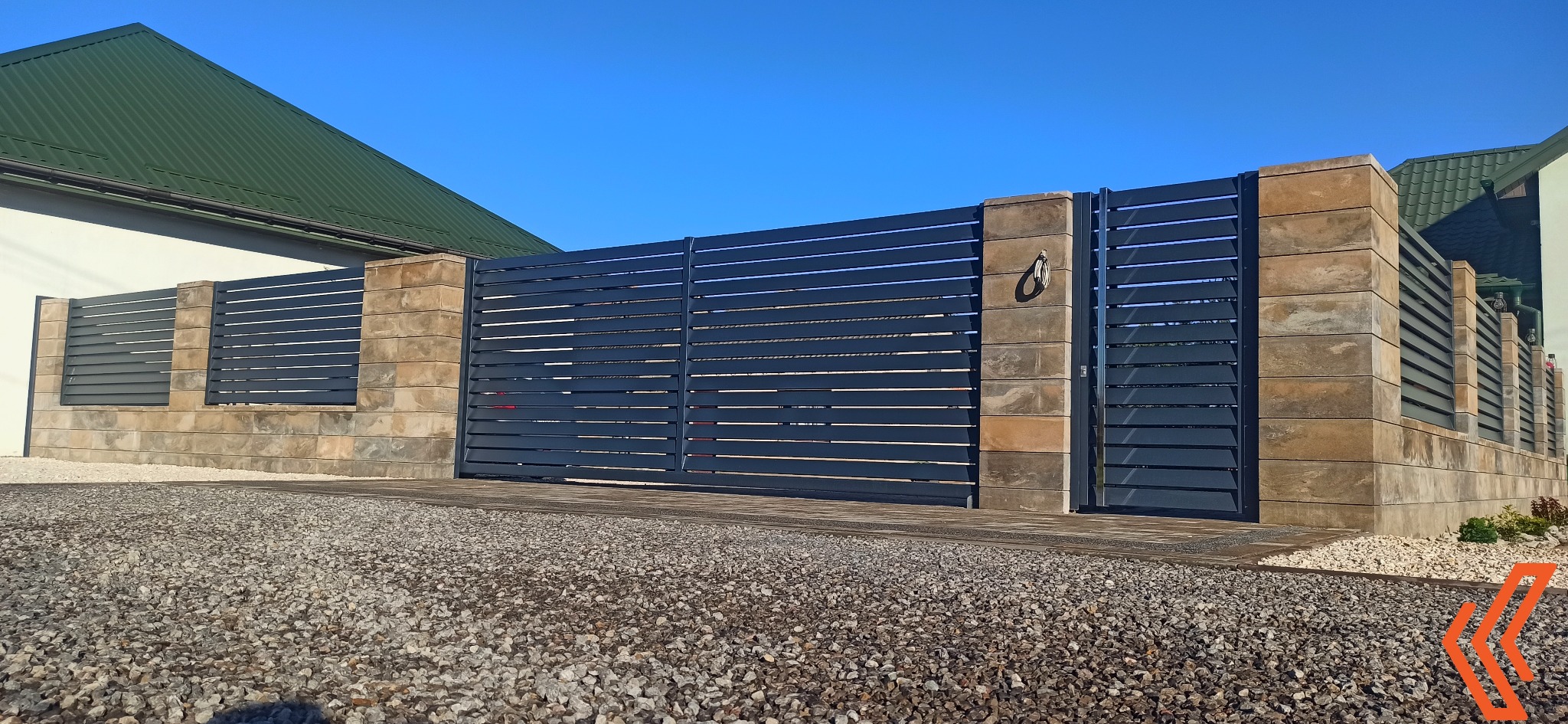 Nowoczesna brama wjazdowa z poziomych, szarych profili, osadzona na słupkach z bloków imitujących kamień, na tle zielonego dachu i błękitnego nieba.