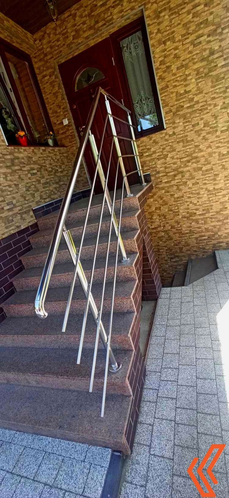 Zewnętrzne schody z brązowego granitu z nowoczesną balustradą ze stali nierdzewnej, prowadzące do ciemnych drzwi wejściowych z szybą i okna z kwiatami w tle, na elewacji z kamienia dekoracyjnego.