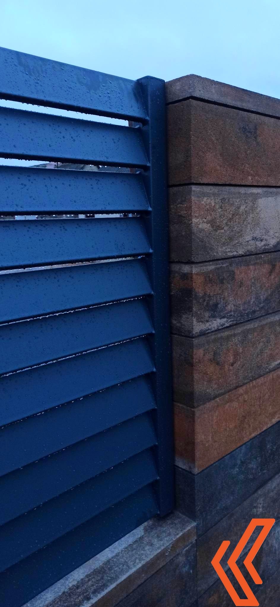 Fragment nowoczesnego ogrodzenia: niebieskie, poziome panele metalowe z kroplami deszczu, połączone z brązowo-szarym słupkiem z bloczków betonowych.