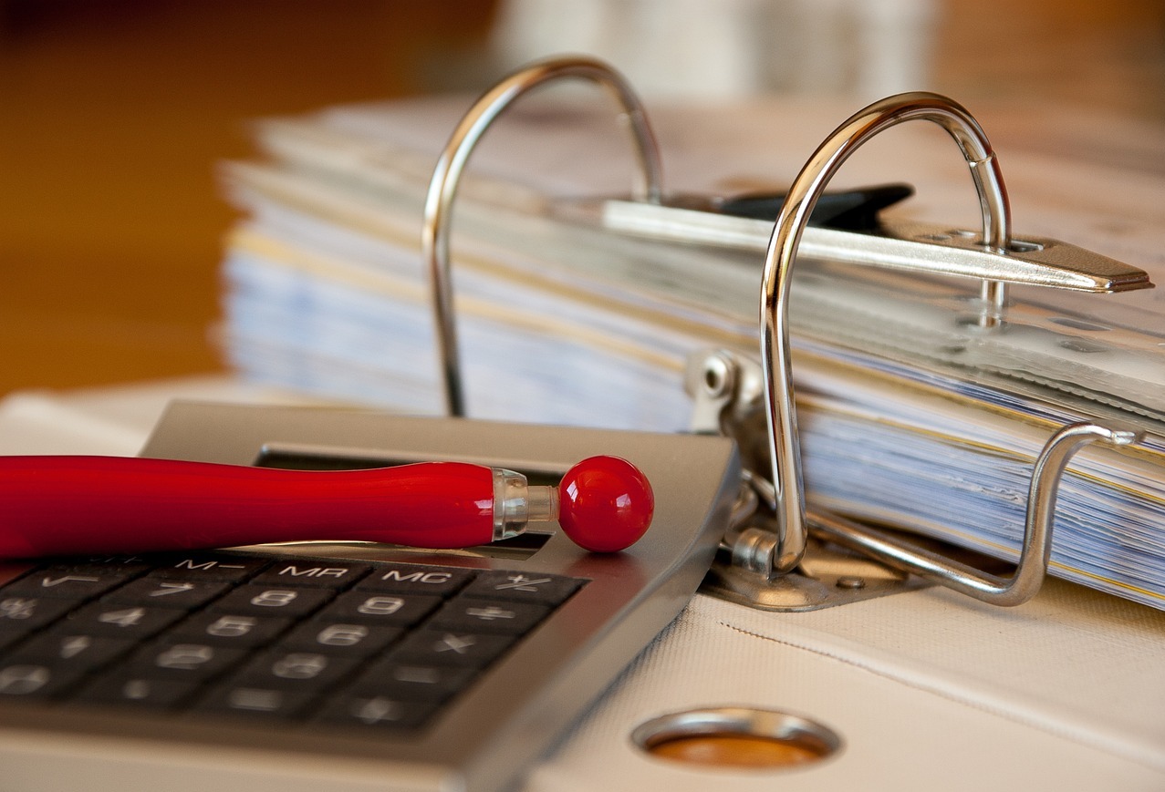 Kalkulator z czerwonym długopisem leżący na segregatorze z dokumentami, widoczne metalowe elementy segregatora.