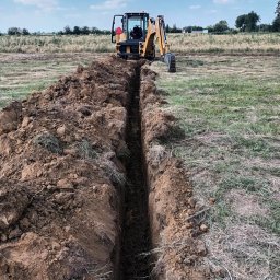 PAT-MAX - Żółta koparko-ładowarka Case stoi na polu za długim, wąskim wykopem w ziemi, z zieloną trawą i suchymi źdźbłami w tle.