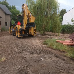 PAT-MAX - Żółta koparko-ładowarka CAT na nierównym, błotnistym terenie z widocznymi śladami opon, w tle domy i drzewa, rury PCV leżą obok małej drewnianej budki.