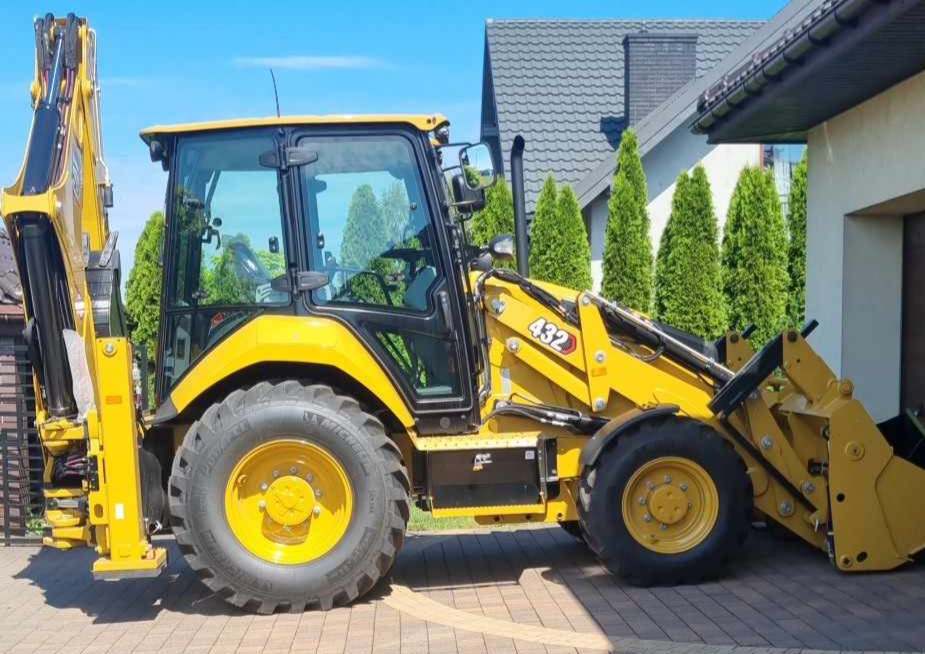 Żółta koparko-ładowarka JCB 432 zaparkowana na kostce brukowej przed domem z szarym dachem i tujami w tle, widok z boku.