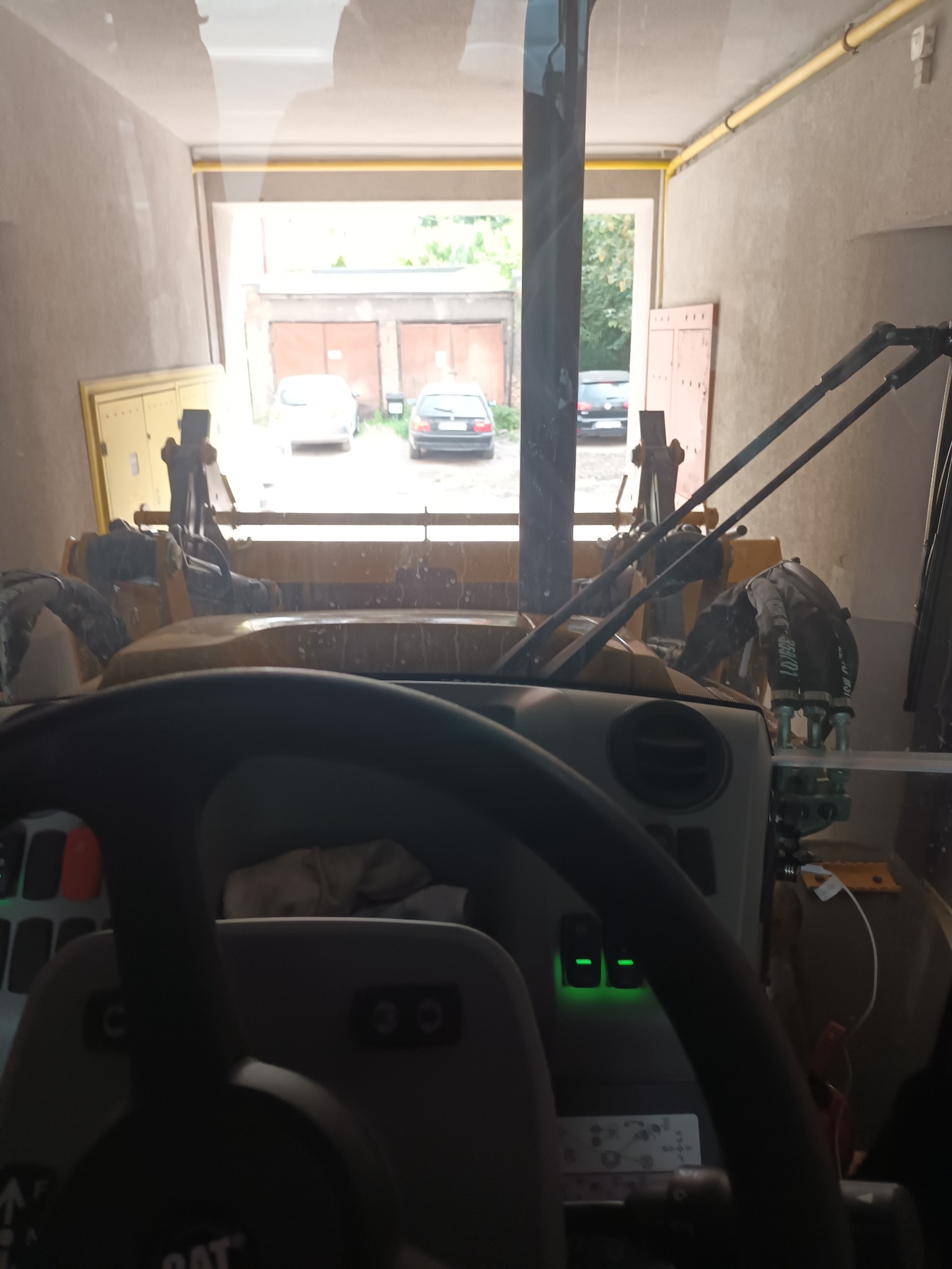 Widok z kabiny ładowarki teleskopowej na plac z zaparkowanymi samochodami, widoczna kierownica z podświetlonymi przyciskami i elementy hydrauliki maszyny.