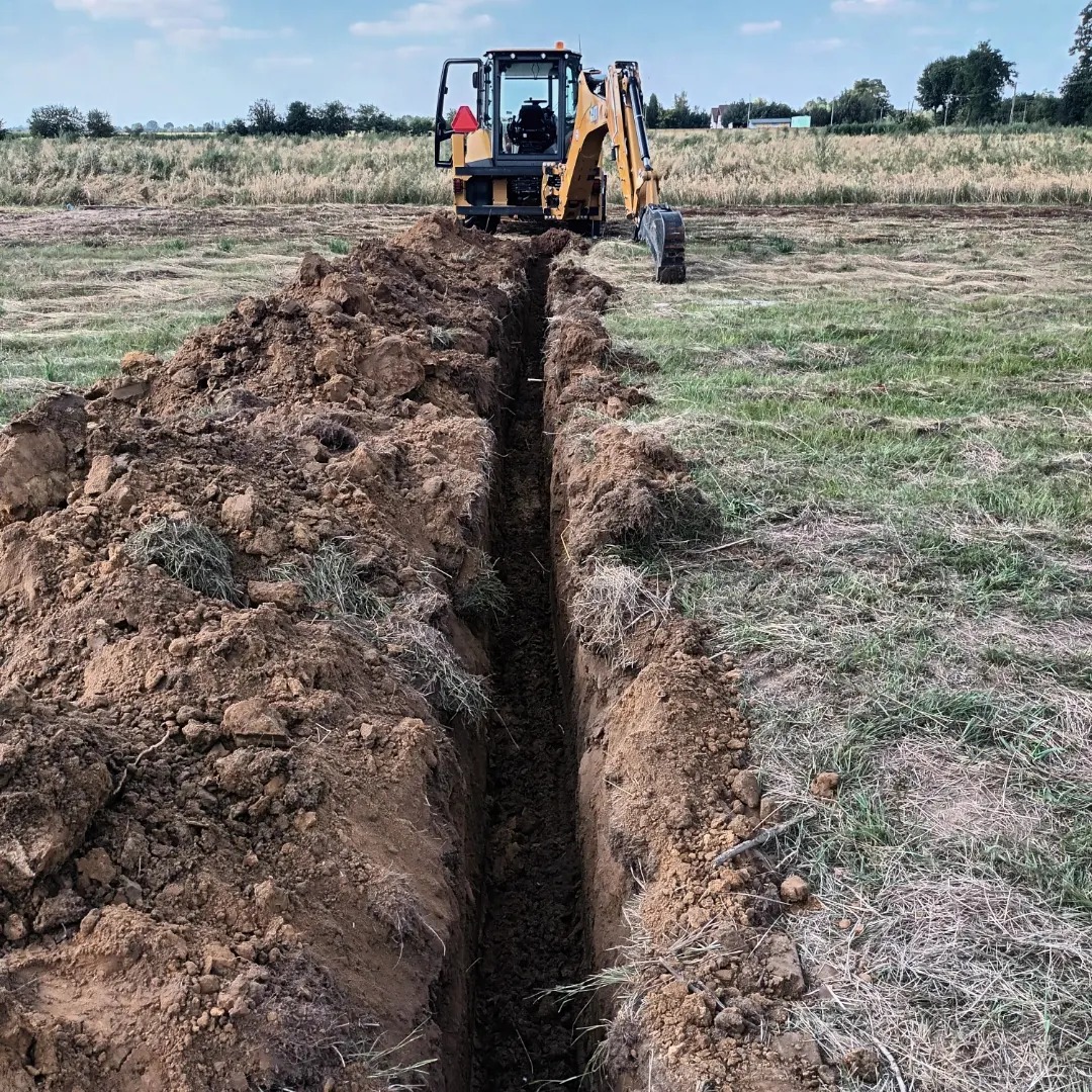 Żółta koparko-ładowarka Case stoi na polu za długim, wąskim wykopem w ziemi, z zieloną trawą i suchymi źdźbłami w tle.