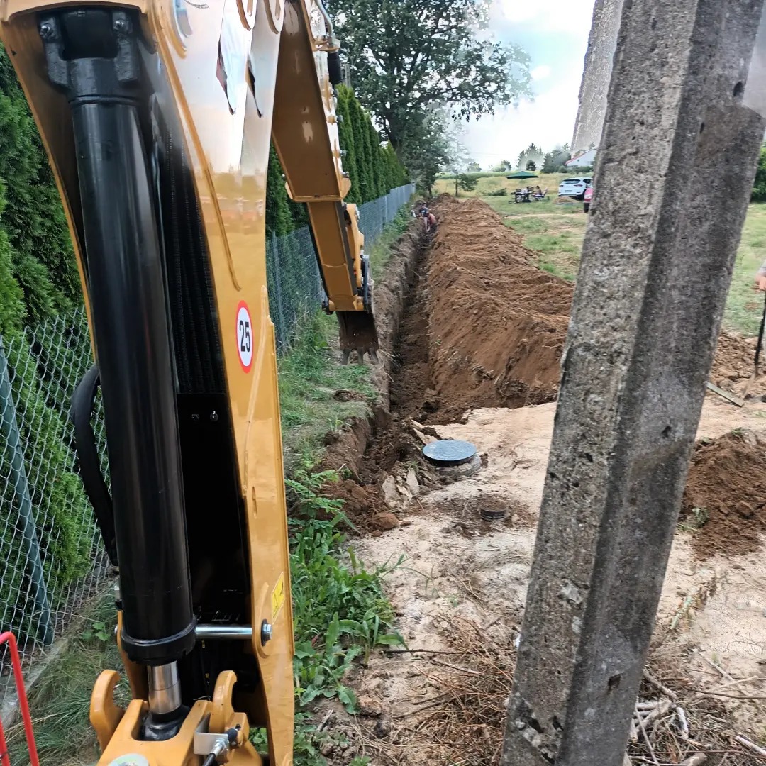 Żółta koparka w trakcie wykonywania wykopu pod instalacje, widoczny rów w ziemi, betonowy słup w pierwszym planie i zielony żywopłot w tle.