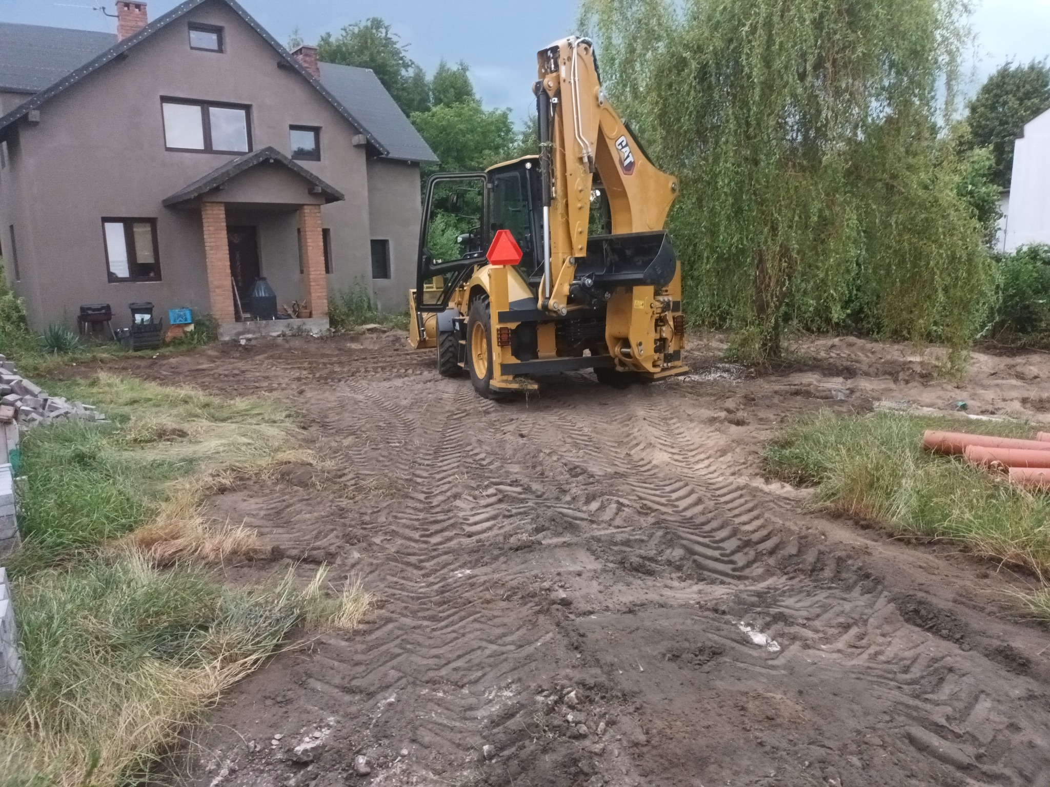 Żółta koparko-ładowarka CAT na terenie budowy przed domem jednorodzinnym, widoczne ślady opon na ubitej ziemi, rury kanalizacyjne i częściowo usunięta darń.