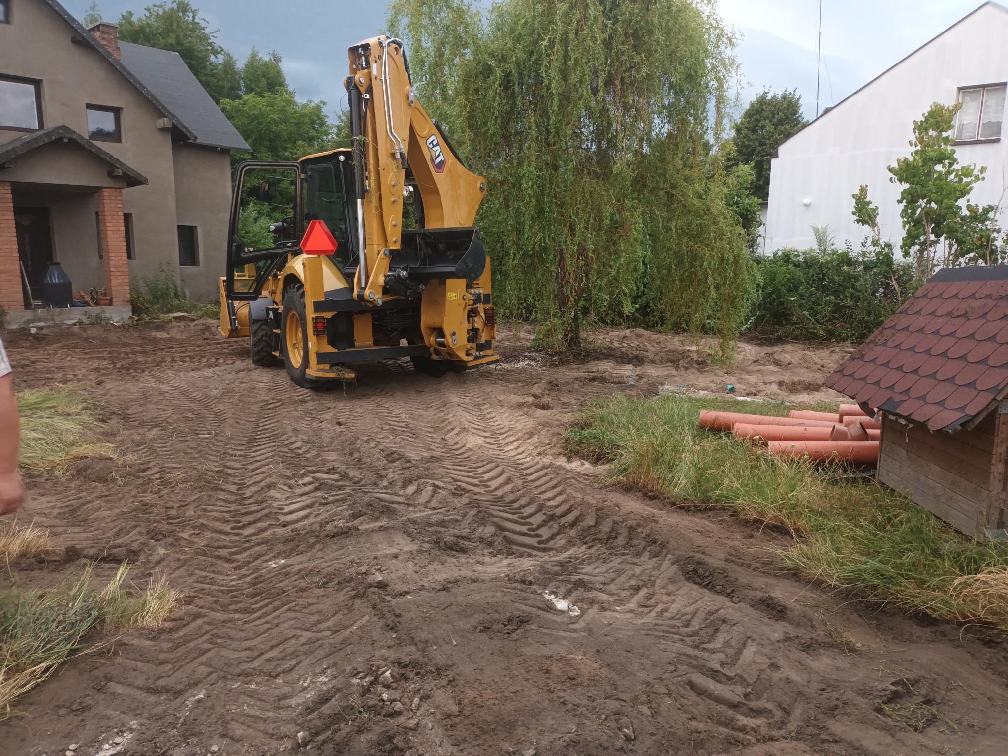 Żółta koparko-ładowarka CAT na nierównym, błotnistym terenie z widocznymi śladami opon, w tle domy i drzewa, rury PCV leżą obok małej drewnianej budki.