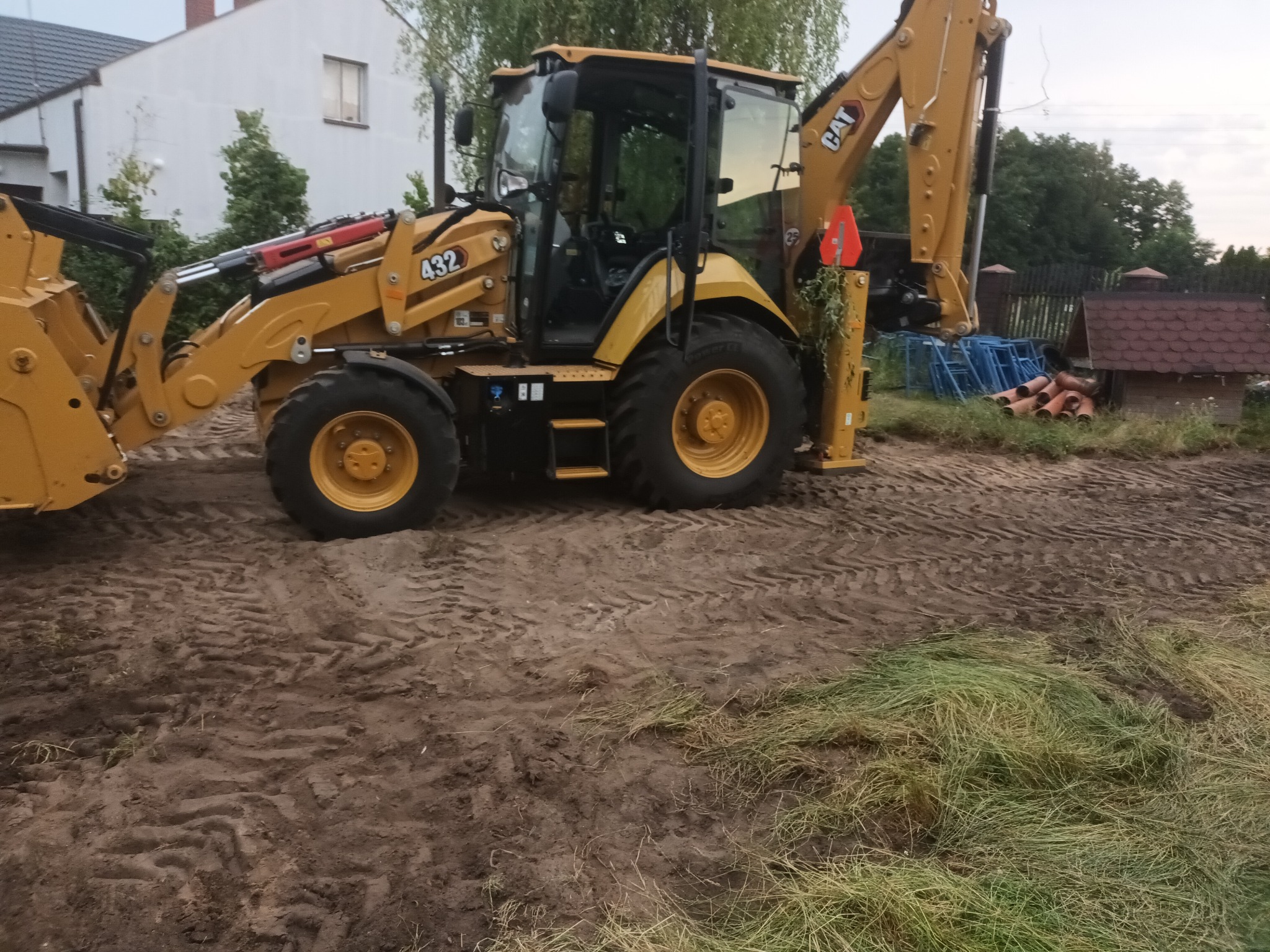 Żółta koparko-ładowarka CAT 432 na nierównym, częściowo zniwelowanym terenie z widocznymi śladami opon, w tle dom jednorodzinny i elementy ogrodzenia.