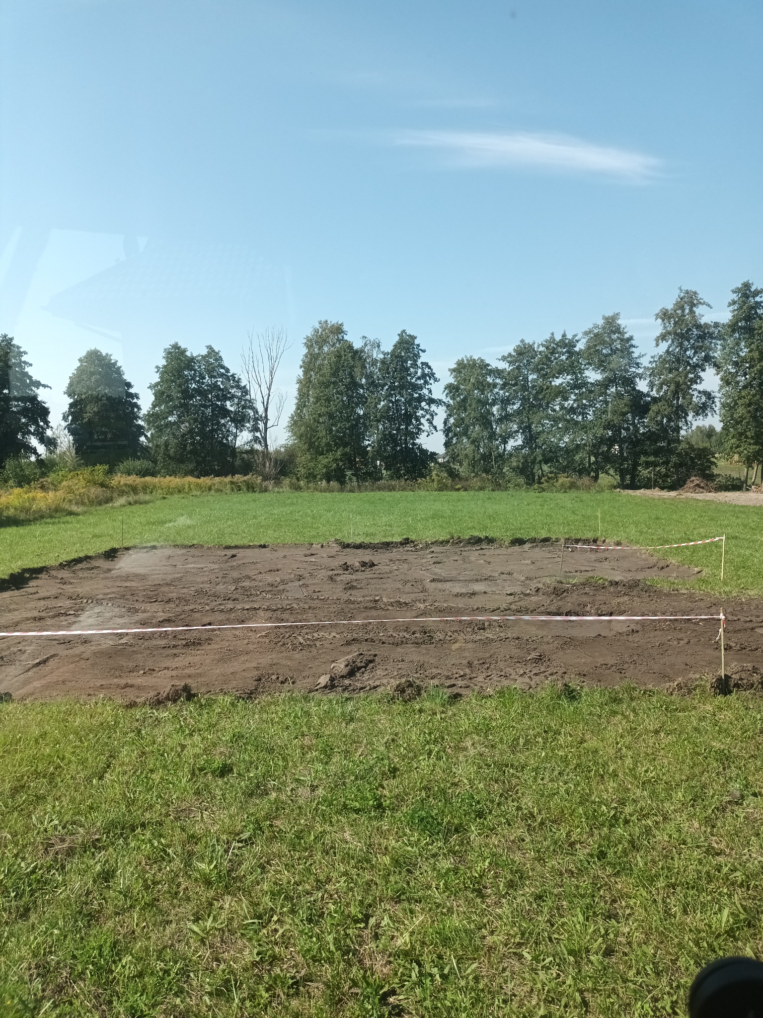 Przygotowany teren pod budowę z wytyczonym obrysem, widoczne słupki i taśmy wyznaczające obszar prac, otoczone zieloną trawą i drzewami w tle pod błękitnym niebem.