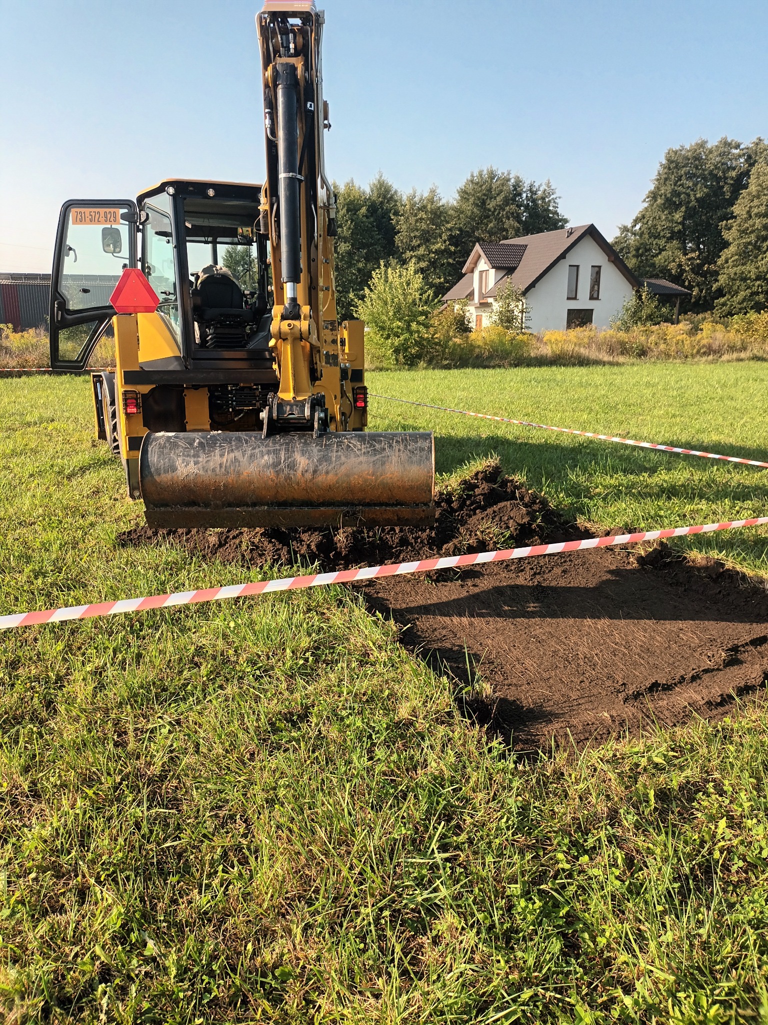 Żółta mini koparka przygotowuje wykop w trawiastym terenie, widoczne biało-czerwone taśmy ostrzegawcze, w tle dom jednorodzinny i drzewa.