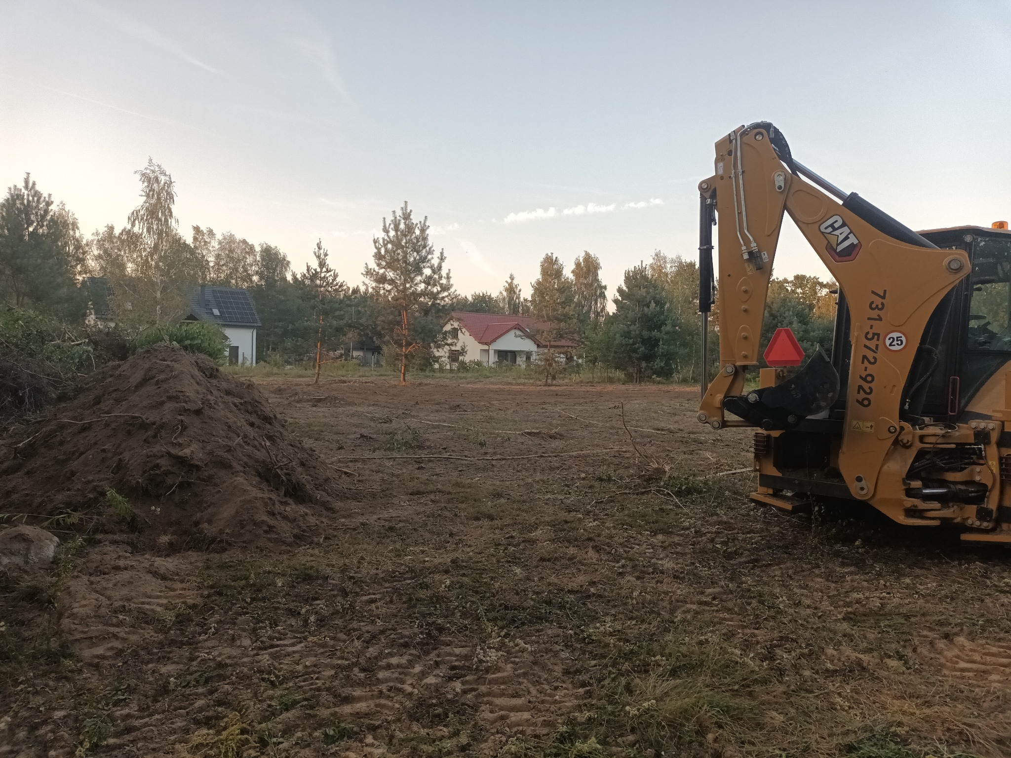 Żółta koparko-ładowarka CAT na działce z usypaną pryzmą ziemi, w tle domy jednorodzinne i drzewa, widoczne ślady opon na gruncie.