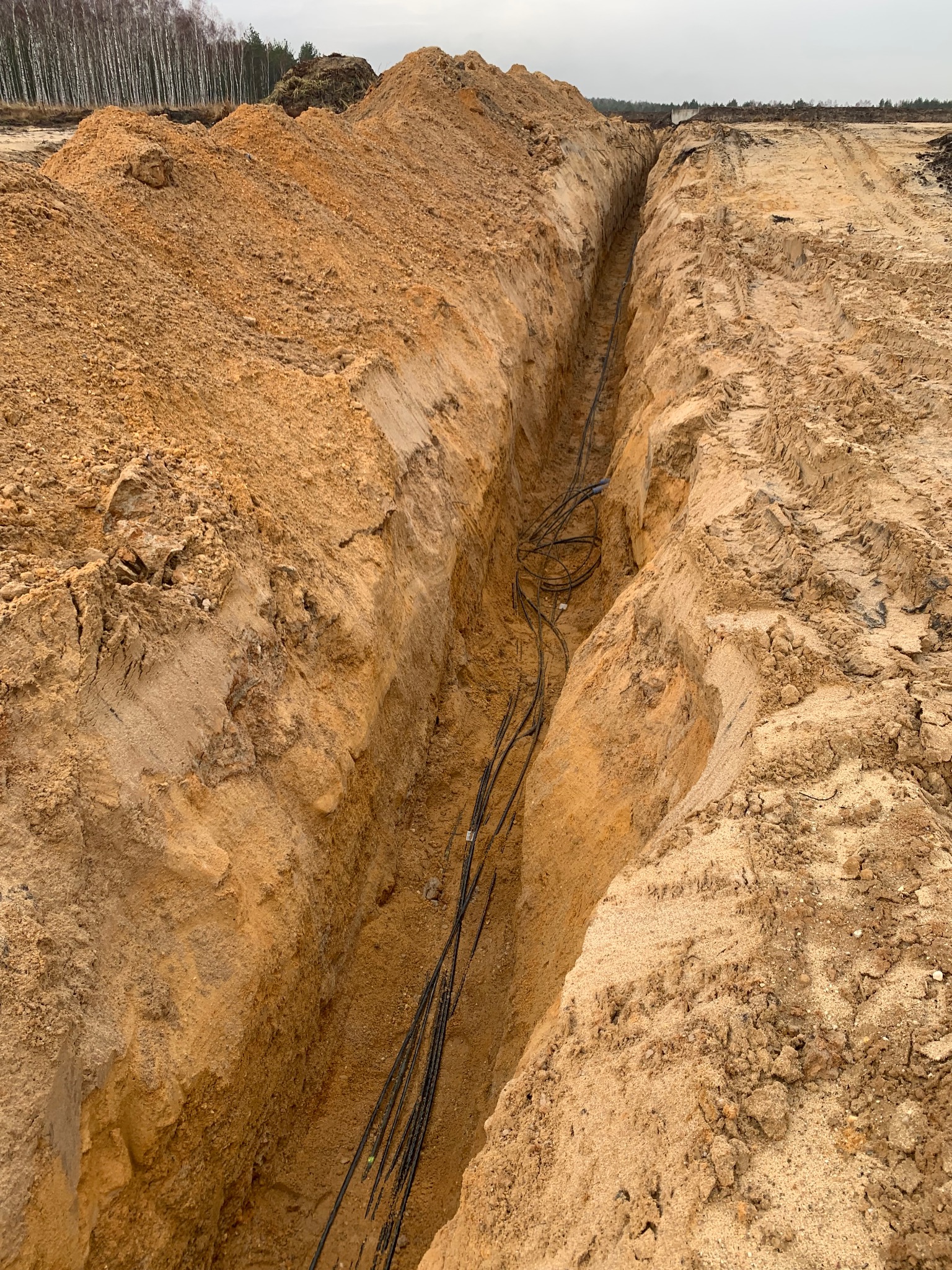 Wąski wykop w piaszczystej ziemi z ułożonymi kablami wewnątrz, widok z góry, ślady opon obok wykopu, na horyzoncie las.