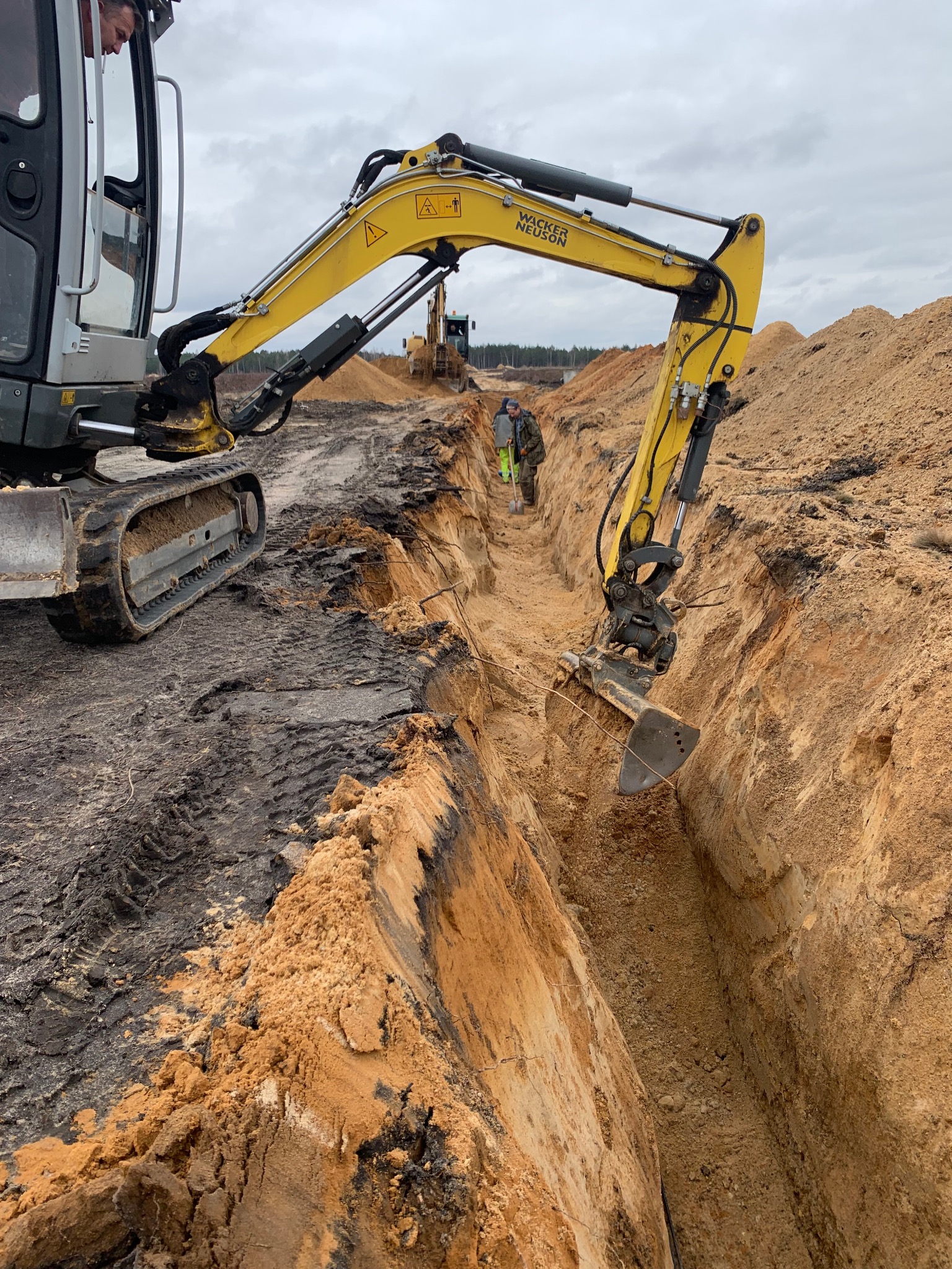 Żółta mini koparka Wacker Neuson pogłębia wąski wykop w piaszczystej ziemi, w tle widoczni dwaj pracownicy z łopatami i druga koparka.