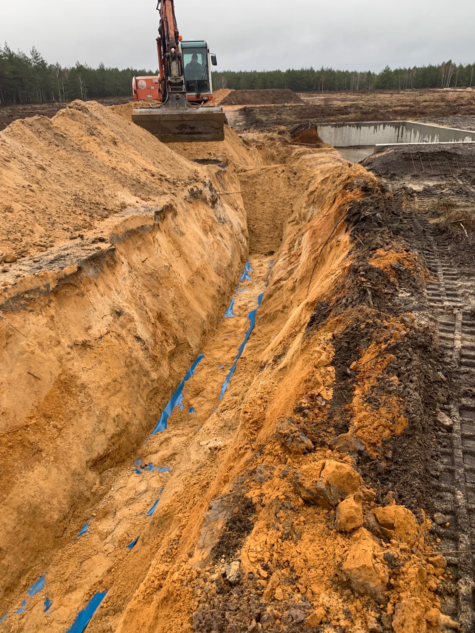 Wykopany rów z niebieską folią na dnie, widoczny z perspektywy góry, z koparką w tle i betonową konstrukcją w oddali na tle lasu