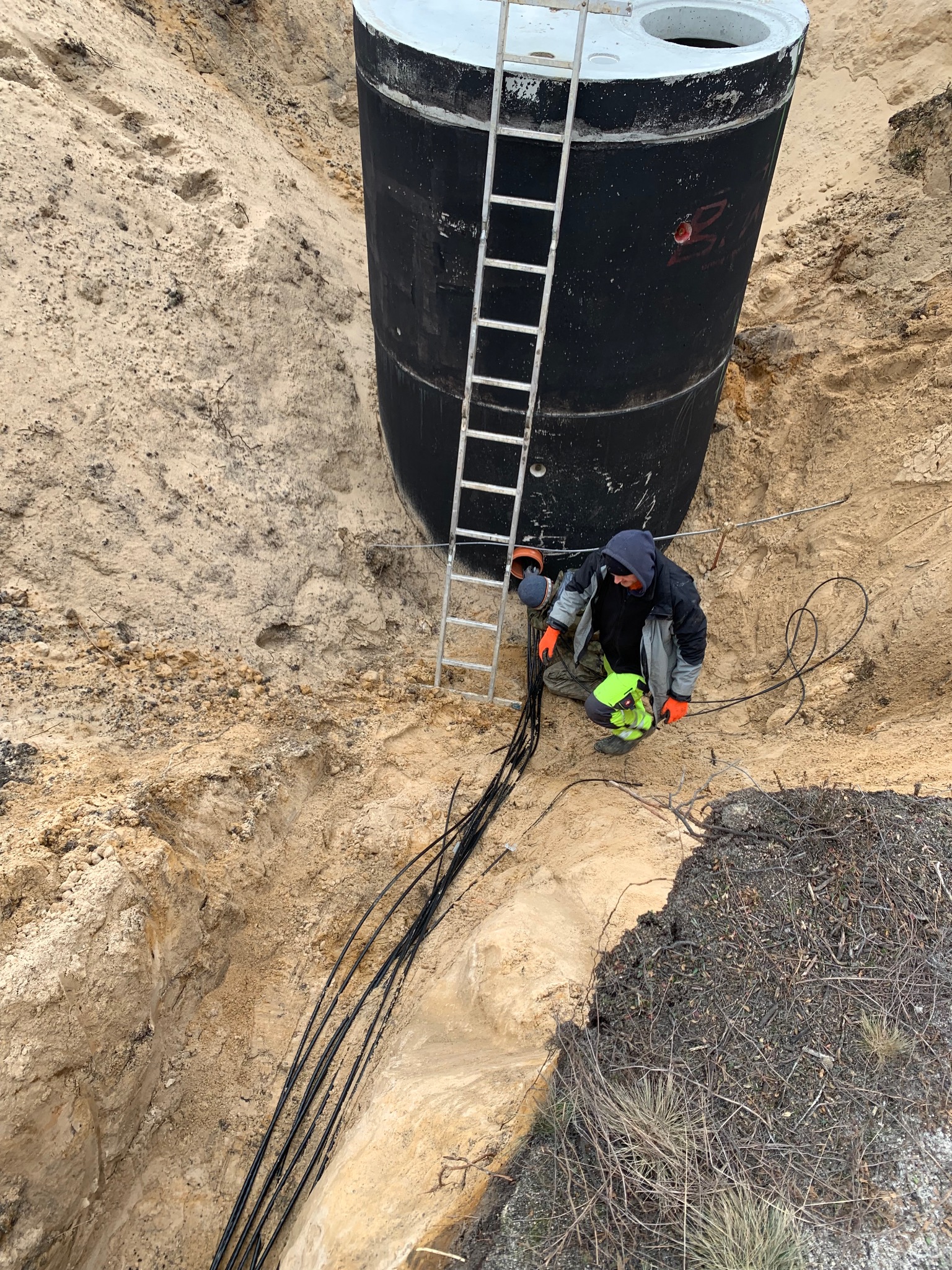 Czarny zbiornik z włazem, umieszczony w głębokim wykopie, z pracownikiem w kasku i rękawicach montującym kable, drabina oparta o zbiornik.